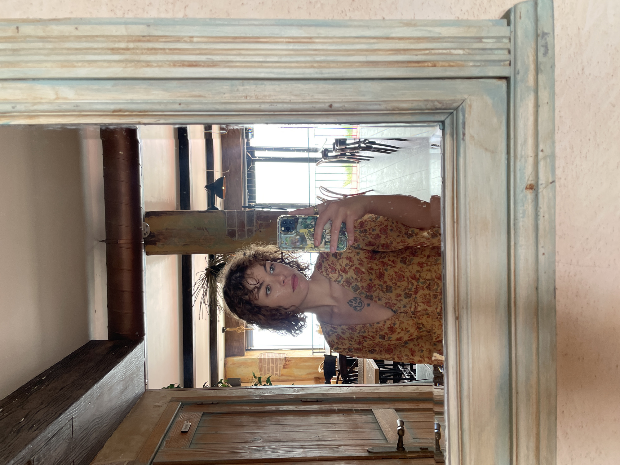 The image is a mirror selfie of a woman with curly hair and a floral dress. She is holding a smartphone with a visible phone case. The reflection shows an interior space with wooden beams, a door, and a window letting in daylight. Hangers can be seen through the window. The mirror has a decorative, slightly distressed frame. There is no definitive information to confirm the location as Rafailovići, Montenegro from the image alone.