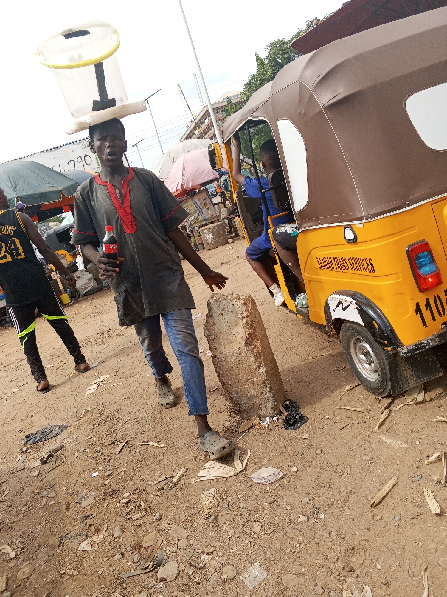 The image captures a street scene in Abuja, Nigeria. A young man is the central figure, walking while balancing a large, transparent container on his head. He holds a Coca-Cola bottle in his left hand and wears a gray shirt with red trim, blue jeans, and gray Crocs. Another man, wearing a black basketball jersey with the number 24, stands nearby. A yellow "keke napep" (auto-rickshaw) is positioned on the right, with a passenger visible inside. The ground is dirt, scattered with litter. Buildings and umbrellas are visible in the background, indicating a market or commercial area.