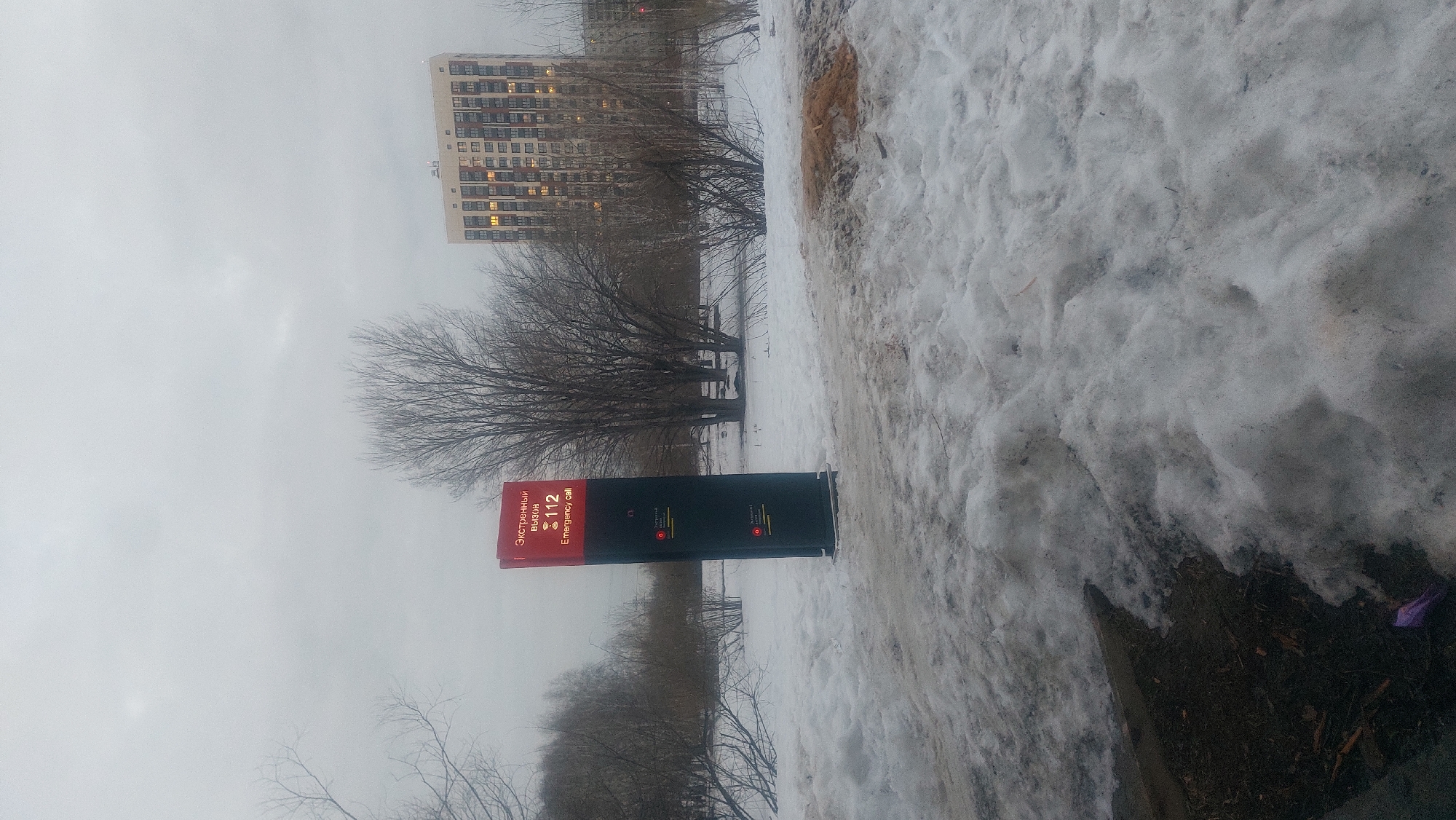 The image shows a snowy scene in Novosibirsk, Russia, possibly in a park or a green area near a residential building. In the foreground, there is an emergency call box with inscriptions in Russian and English reading "Emergency call 112". Bare trees are visible behind the call box. A tall, multi-story residential building stands in the background. The ground is covered with snow, which appears to be melting in some areas. The sky is overcast, indicating a dull weather.