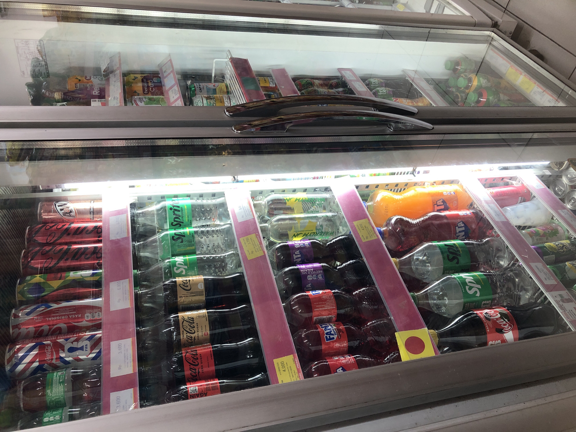 This is a medium shot, eye-level, indoor photograph of a brightly lit beverage refrigerator display. The refrigerator, located in Putat, Indonesia, is filled with various bottled and canned drinks. The left side of the refrigerator is filled with soda bottles, primarily Coca-Cola and Fanta, in red, orange, and clear plastic bottles. There are also several bottles of Sprite, a clear carbonated beverage, with green labeling. Above the soda bottles, on a top shelf, are several cans of Coca-Cola, with red and white designs, and other soda cans with various colorful designs. The right side of the refrigerator contains more canned drinks, including what appears to be Fanta and other brands with labels in Indonesian. The refrigerator doors are glass, allowing a clear view of the products inside. The overall environment appears to be a small convenience store or a similar retail establishment.