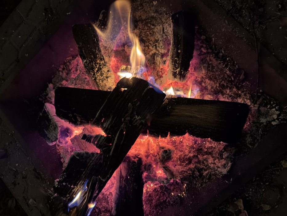 The image displays an overhead, close-up view of an active fire contained within a dark, square or rectangular fire pit, located in Huixquilucan de Degollado, Mexico. Multiple pieces of wood, appearing as dark, irregularly shaped logs, are in various stages of combustion. The core of the fire consists of intensely glowing red and orange embers, partially covered by white and grey ash. Vibrant flames, primarily yellow and orange, with distinct blue streaks indicating higher temperatures, emanate from and around the burning wood, particularly visible on the left side and extending towards the center-right. No individuals or other discernible objects are present within the immediate scene. The environment outside the fire pit is obscured by darkness, consistent with nighttime conditions.