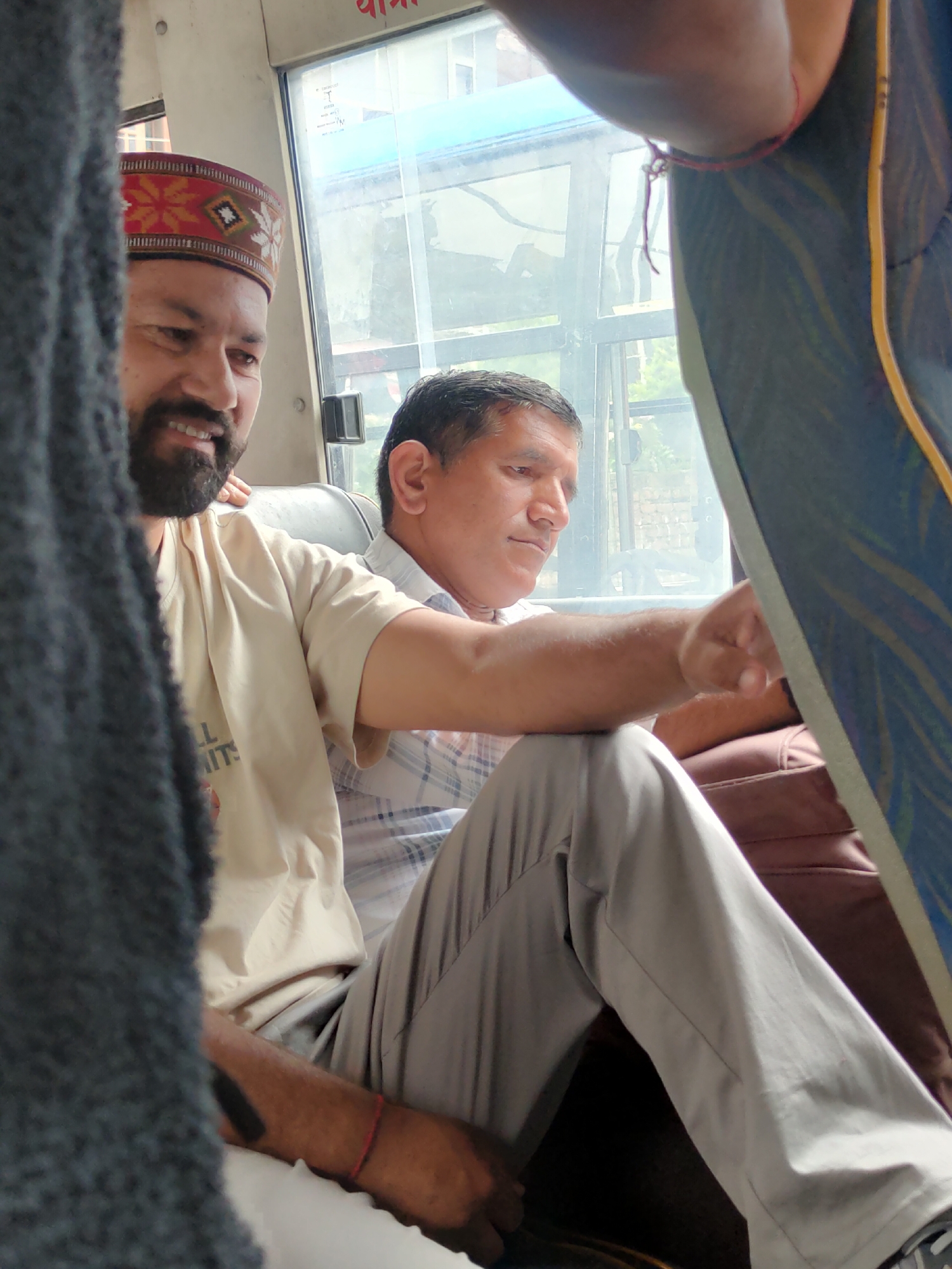 The image shows two men seated inside what appears to be a bus or a vehicle, likely in Manali, India, given the context of the location. The man on the left is wearing a traditional Himachali cap, which is red with intricate embroidered patterns of yellow, black, and white. He has a beard and is smiling, with his head turned to the left. He is wearing a light-colored t-shirt with some text that is partially obscured.
The man on the right is wearing a plaid shirt and grey trousers. He is looking to his right, away from the camera. Both men are seated next to each other, suggesting they are traveling together or are passengers in the same vehicle.
The interior of the vehicle is visible, with windows showing a blurred outdoor scene, possibly of trees and buildings. The seating appears to be upholstered in a dark fabric. A part of a dark, textured object, possibly a scarf or shawl, is in the foreground on the left side of the image, partially obscuring the man on the left. A red string bracelet is visible on the right wrist of the man on the left. The overall lighting suggests it is daytime.