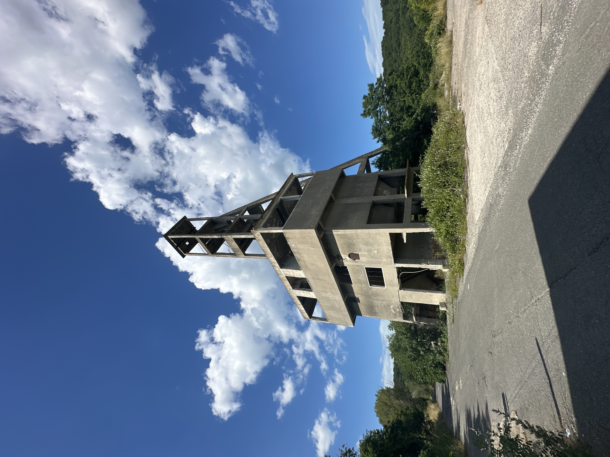 An abandoned concrete structure, likely a mine shaft headframe, stands under a bright blue sky with scattered white clouds. The structure is multi-tiered and appears to be in a state of disrepair, with open doorways and missing sections. Vegetation, including trees and bushes, surrounds the base of the structure and encroaches on a cracked asphalt road leading away from it. The scene is situated in a rural or semi-rural setting, with a wooded hillside visible in the background. The lighting suggests it is daytime. There are no people or animals present in the image. The location is identified as Rosen, Bulgaria.