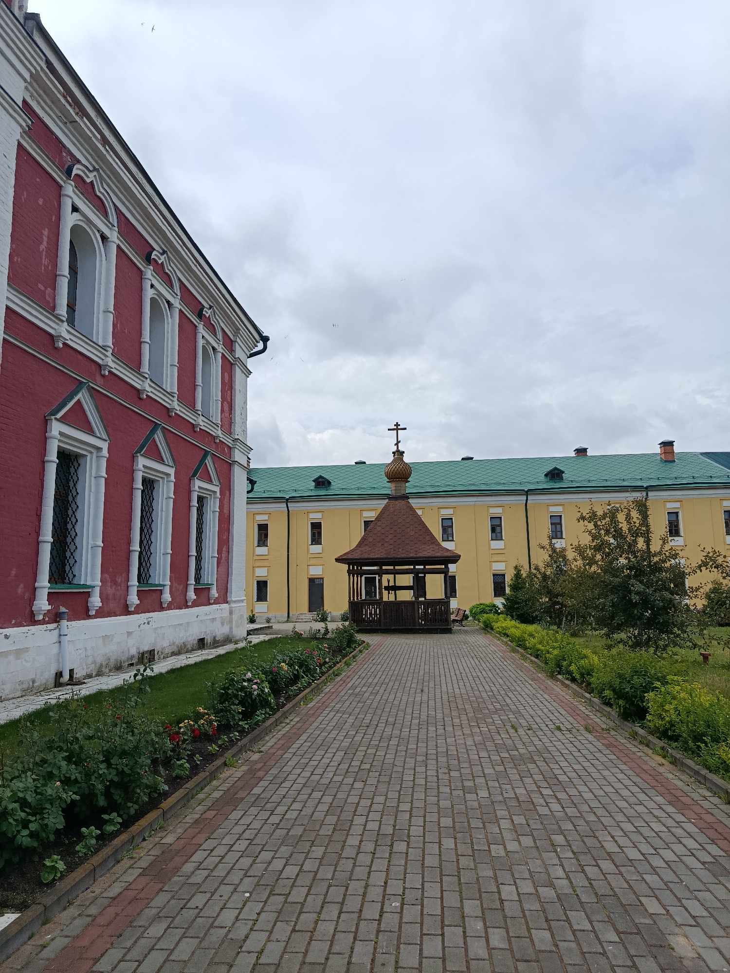 The image depicts an exterior view of the Svyato-Uspensky monastery in the village of Radovitsy, Russia. On the left, a portion of a large red brick building with white trim and arched windows is visible. In the center, a pathway paved with gray and red bricks leads towards a brown wooden gazebo with a domed roof and a cross on top. Behind the gazebo, a long, yellow building with a green roof and numerous windows stretches across the frame. The foreground features a flowerbed with roses and green grass. The sky is overcast with clouds. There are no people visible in the image.