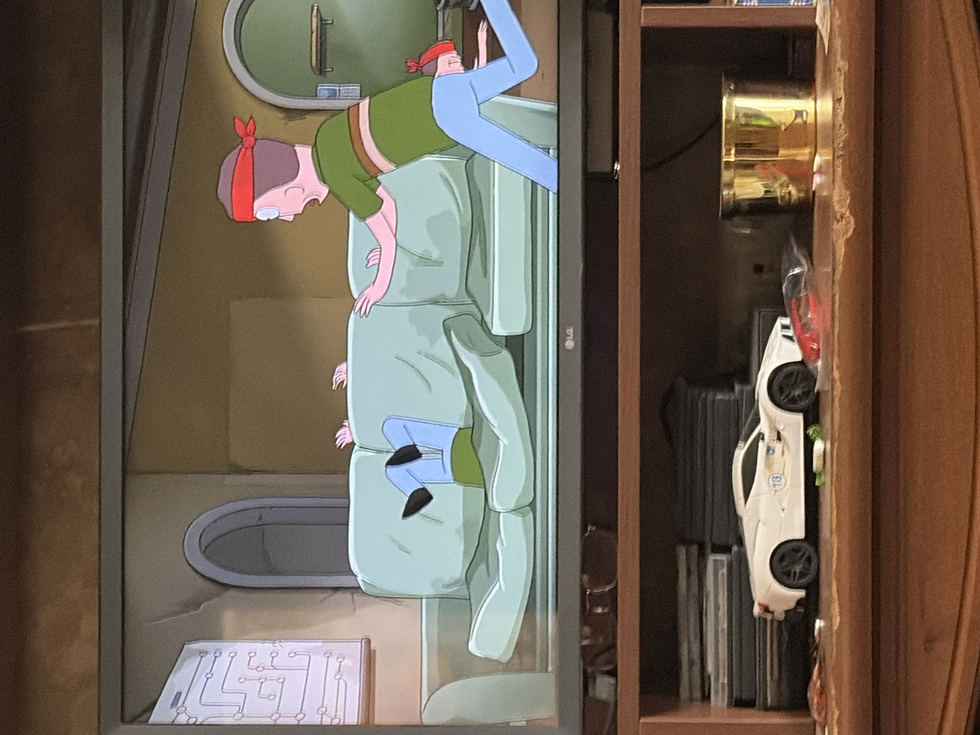 The image shows a television screen displaying an animated scene and the shelf below it. On the television screen, two animated characters are visible. One character, wearing a red headband and a green shirt with brown stripes, is reaching out towards a light blue couch. Another character, wearing blue pants and a green shirt, has their legs and torso on the couch, with their head and arms unseen. The background on the screen features a circuit board diagram on a wall, an arched doorway, and a plain wall. The shelf below the television holds several items. A white toy car with a racing decal is positioned on the shelf. To the left of the car, stacks of what appear to be DVDs or similar media are visible. To the right of the car, a golden cylindrical container is present. The shelf is made of wood and shows some wear and tear. The overall scene depicts a living room entertainment setup with a focus on the television content and the display shelf.