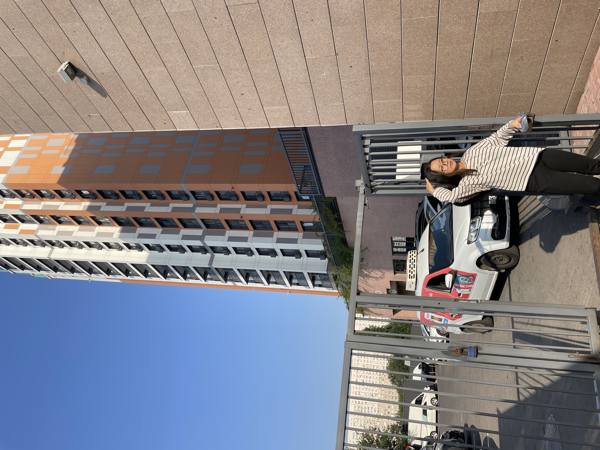 In Almaty, Kazakhstan, a young woman with dark hair and glasses, wearing a striped sweater and black pants, stands by a grey metal gate, her left hand touching her hair and her right hand holding a clear plastic bag. Behind her, a white van with red and white logos is visible, along with other vehicles and a tall, modern apartment building with orange and grey panels. The surrounding environment includes a textured brown wall of a building to the right, and a clear blue sky above.