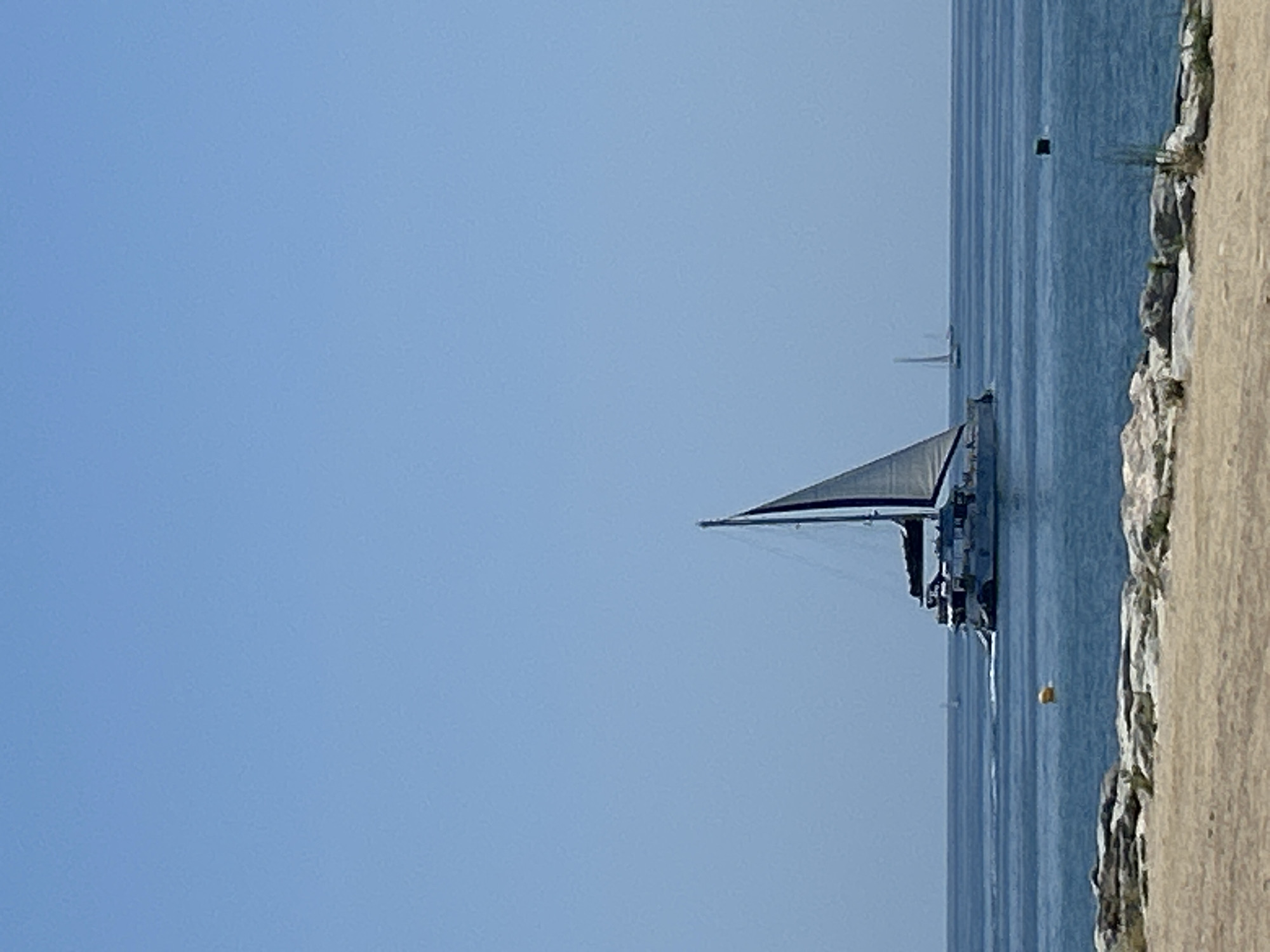 A wide-angle, eye-level shot captures a bright, clear day on the Mediterranean Sea, with a Catalan coastline visible in the foreground. The sky is a uniform, cloudless blue, stretching across the entire upper half of the frame. The sea is a deep, calm blue, with gentle ripples reflecting the sunlight.
Dominating the mid-ground is a large, white catamaran with two masts, its sails partially unfurled. The vessel appears to be in motion, cruising across the water. A smaller white sailboat is visible in the distance, further out at sea. A yellow buoy floats near the center of the water.
The foreground consists of a rocky, sandy shoreline. Large, greyish-brown rocks are scattered along the edge of the water, with dry, sandy ground extending into the lower part of the frame. The image was taken in Barcelona, Spain.