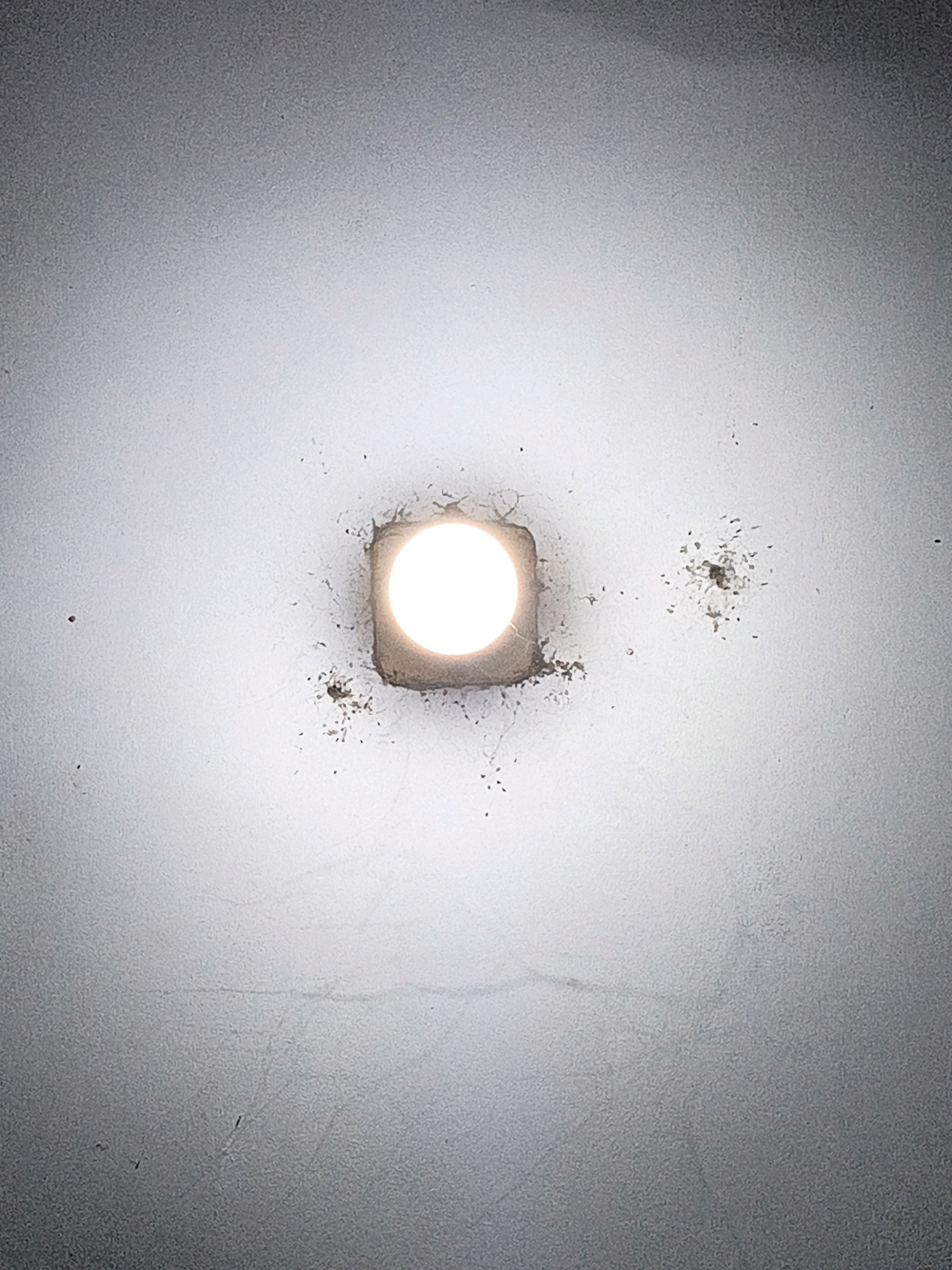 An overhead, close-up, medium-shot photograph captures a circular light fixture on a white ceiling in West Jakarta, Indonesia. The light emits a bright white glow, creating a halo effect around the fixture. The ceiling surface appears textured and shows signs of age, with some discoloration and faint cracks visible. Small, dark specks, possibly dust or debris, are scattered across the ceiling, particularly concentrated around the light fixture. The overall lighting is dim, with the light fixture being the primary source of illumination. The scene is static and lacks any human presence or activity.