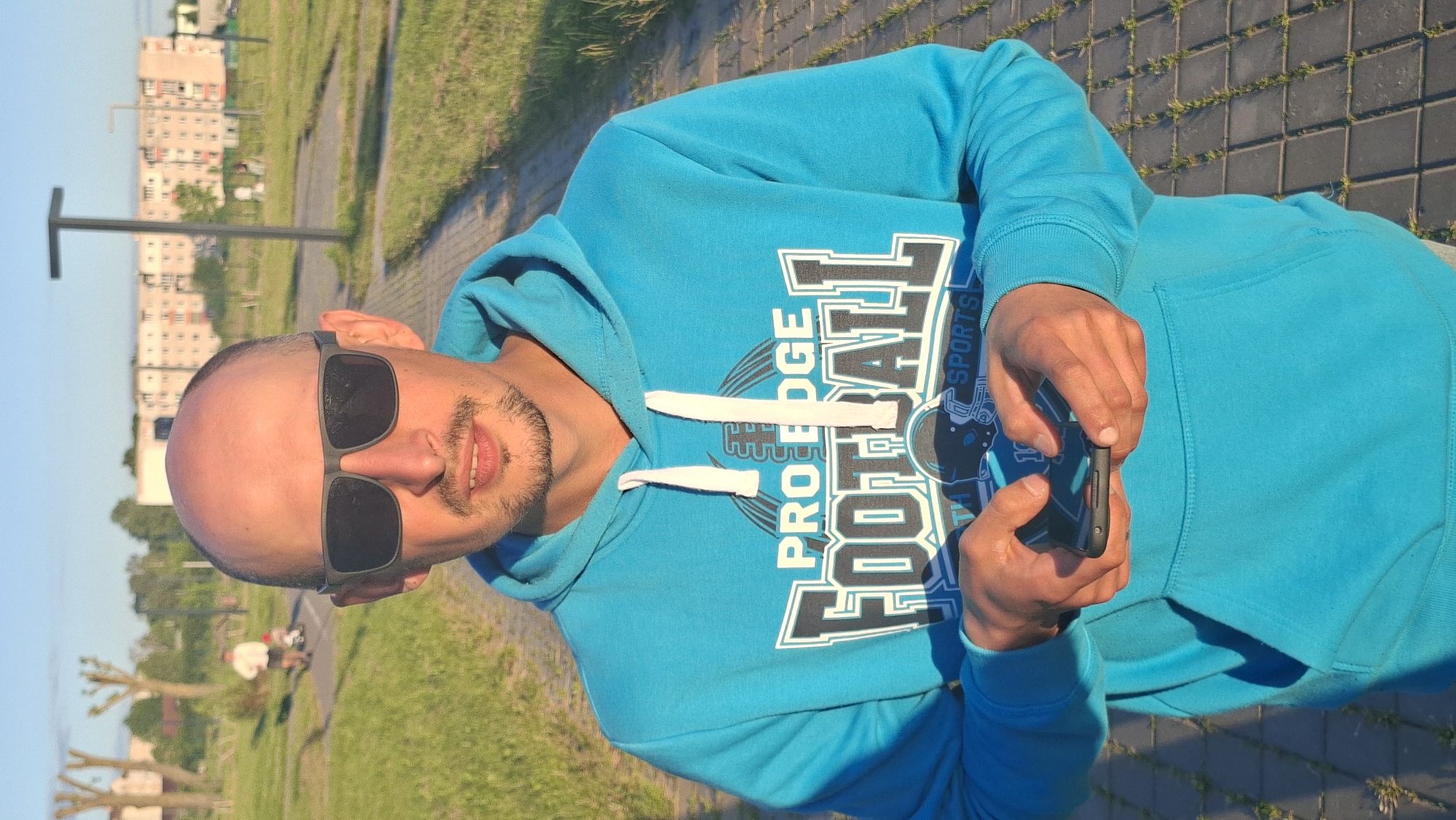 In the city of Smaliavichy, Belarus, a man with a shaved head, stubble, and sunglasses stands on a paved path, holding a smartphone in his hands. He is wearing a bright blue hooded sweatshirt with the words "PRO EDGE FOOTBALL" and "SPORTS" printed on it. The background shows a park-like setting with a paved walkway, green grass, and trees. In the distance, there are residential buildings and a lamppost. The sun is shining, casting long shadows.