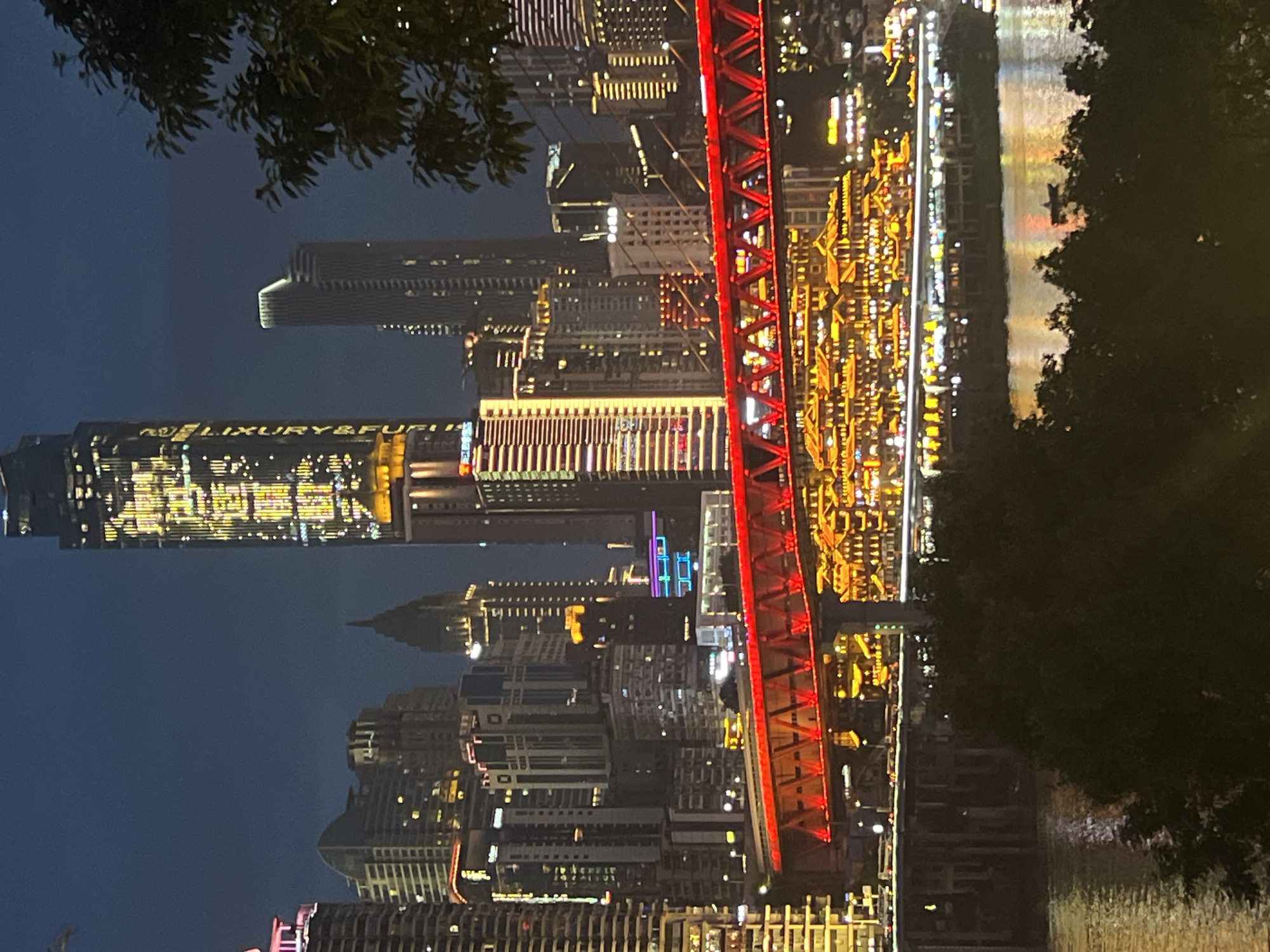 This media file is a nighttime photograph of a cityscape, likely taken from across a body of water. The image is oriented vertically, with the top of the buildings appearing at the bottom of the frame and vice versa.
The scene is dominated by a dense collection of illuminated skyscrapers and other multi-story buildings. Many of these structures are adorned with numerous lights, creating a vibrant and dazzling urban landscape. A prominent feature of the image is a large red truss bridge spanning across the scene, its structure also lit up. The lights from the buildings and the bridge reflect on the water in the foreground. A small boat with a single silhouetted figure can be seen on the water. Lush green trees are visible in the upper left and right portions of the frame, framing the city view.
The time of day is clearly night, as indicated by the dark sky and the extensive use of artificial lighting. The weather appears to be clear, allowing for good visibility of the city lights and their reflections. The presence of the bridge and the architectural style of the buildings suggest an urban environment, consistent with the provided location context of Jiangbei City Street, China. No specific activity or event is explicitly captured, but the illuminated city suggests a bustling urban environment.
The word "LUXURY&FURNITURE" is visible in large, illuminated lettering on one of the buildings. The overall mood conveyed is one of urban vibrancy and nighttime beauty.