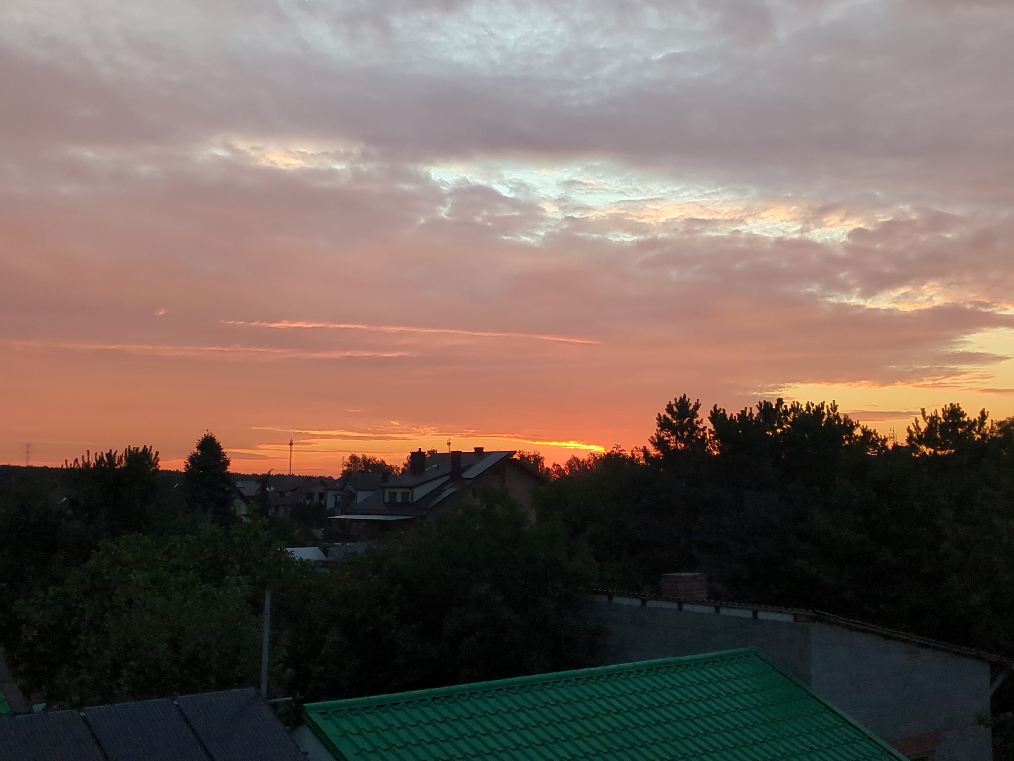 This image depicts a residential area in Rusiec, Poland, during a sunset. The sky is filled with hues of orange, pink, and white, with scattered clouds. Silhouetted against the vibrant sky are trees, rooftops of houses, and a communication tower in the distance. The foreground shows parts of residential rooftops, one with a green tiled roof and another with dark grey panels, suggesting a suburban or semi-rural setting. The overall impression is a tranquil evening scene over a Polish town.