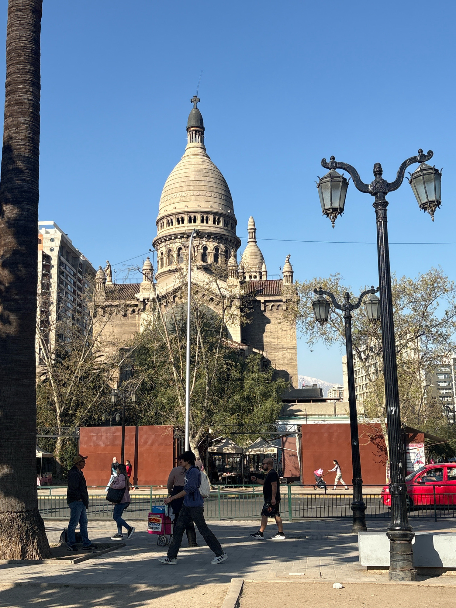 This is an exterior, eye-level, daylight, medium shot of the Metropolitan Cathedral of Santiago, Chile, taken from across a street. The cathedral's dome and parts of its facade, constructed from light-colored stone, are visible. A tall palm tree trunk partially obscures the left side of the image. In the foreground, several people are walking on a sidewalk. One man in a hat and blue jeans is facing left, talking to a woman in a pink jacket and jeans who is looking forward. Another man in a dark shirt and shorts is walking away from the camera, and a person in a blue hoodie and dark pants is pushing a cart with items. A red car is parked on the street on the right side of the image. Streetlights with multiple globes stand on either side of the foreground. In the background, behind the cathedral, is a modern building with many windows. The sky is clear and blue.