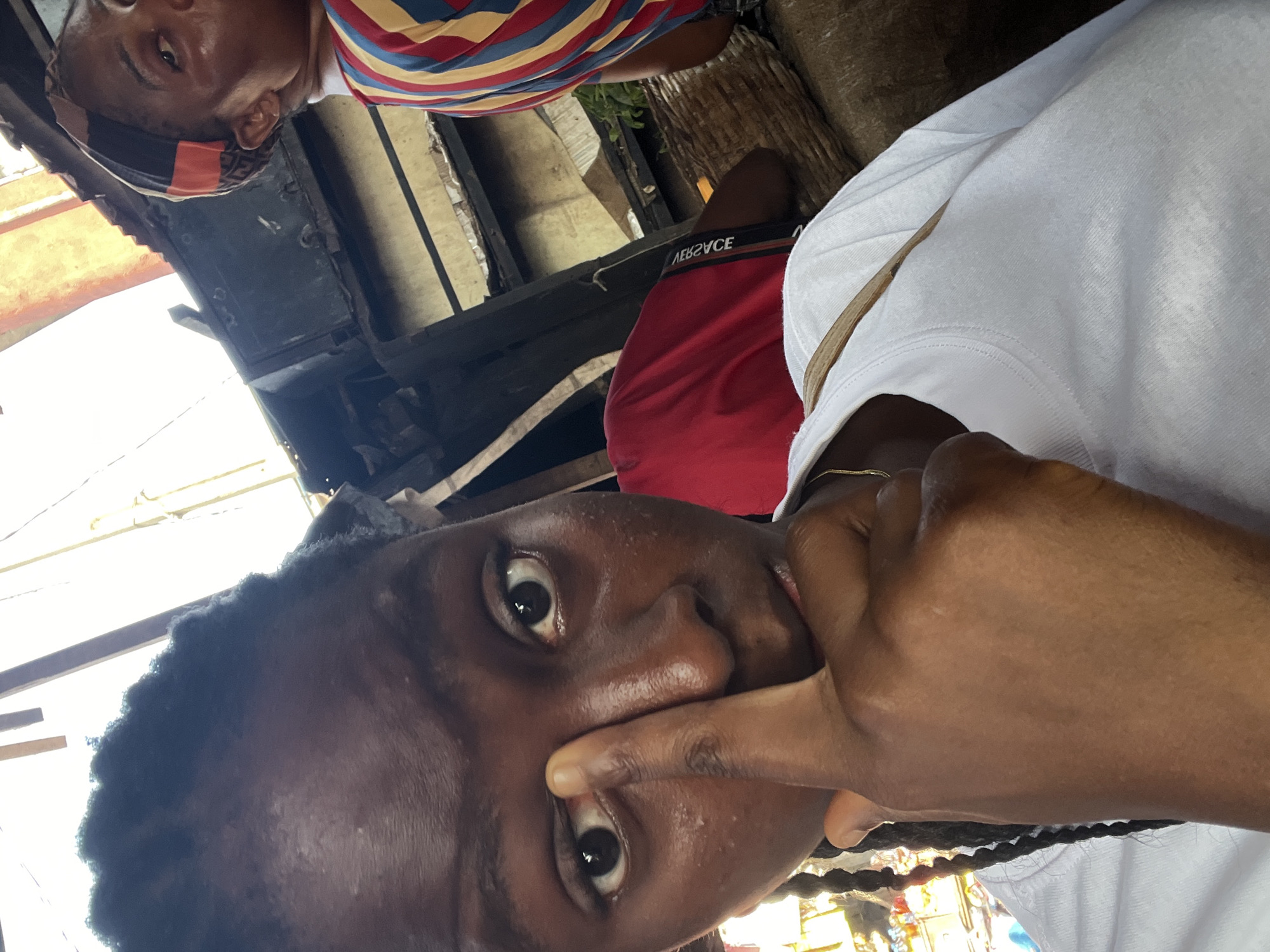 A close-up, selfie-style shot captures a young Black person's face, partially obscured by their right index finger pressed against their eye. Their dark braids are visible on the left side of their face, and they wear a white t-shirt. The background is blurry and appears to be an outdoor market stall, possibly in Nkpor, Nigeria, with hints of wooden structures and other people in the distance. One individual in the background wears a striped shirt, and another is seen in a red shirt. The lighting suggests it is daytime.