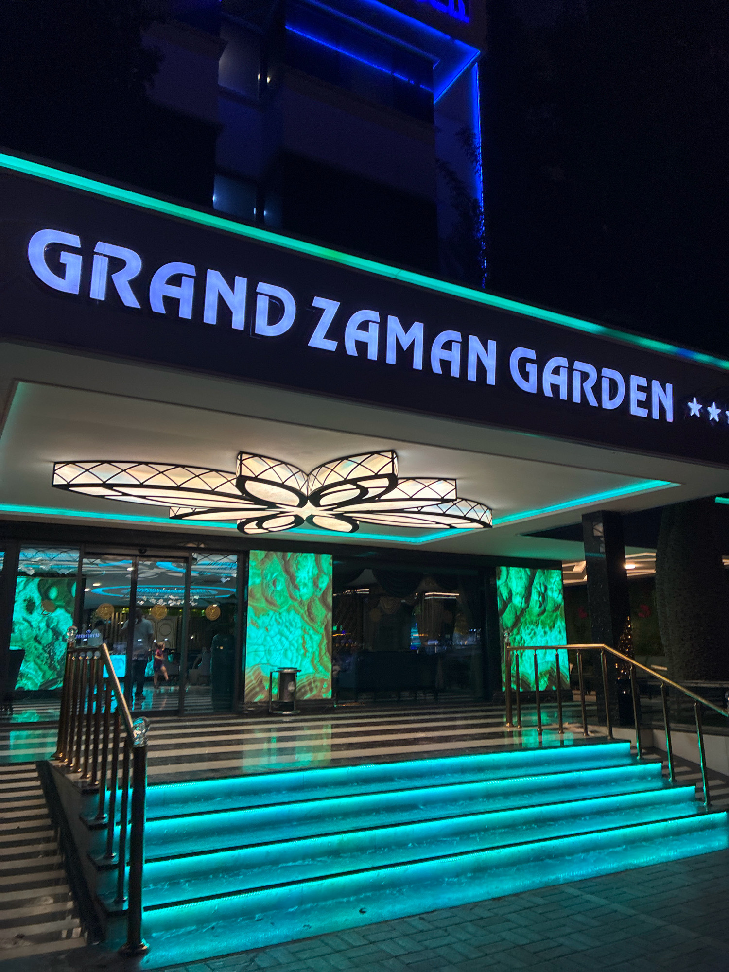 A nighttime exterior shot captures the entrance to "GRAND ZAMAN GARDEN" in Alanya, Turkey. The hotel's name is illuminated in large, white, sans-serif font against a dark background, bordered by a vibrant green neon light. Above the entrance, a modern, multi-tiered light fixture with a geometric pattern is visible. The building's architecture features blue neon lighting accents.
The entrance itself is a set of wide stairs leading up to a glass-doored entryway. The stairs are illuminated from beneath with bright turquoise LED lights, creating a striking visual effect. The steps have a dark and white striped pattern. A metallic railing with decorative posts guides visitors up the stairs. Inside the lobby, through the glass doors, digital screens displaying a green, abstract pattern are visible, along with a few people. The ground outside the entrance is paved with cobblestones. The overall scene suggests a lively and modern hotel entrance, well-lit and inviting for guests.