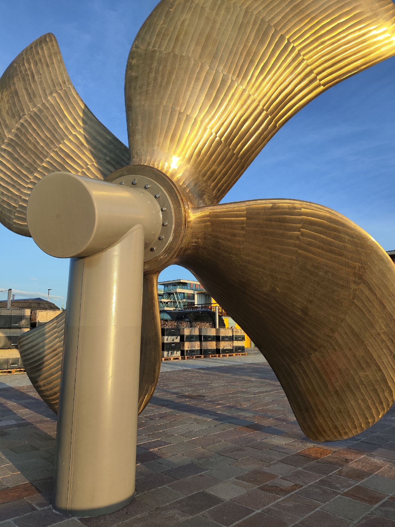 The image shows a large, golden propeller sculpture, likely on display outdoors. The propeller is positioned in the foreground, with its blades angled upwards and outwards. The surface of the propeller exhibits a textured, ribbed pattern, catching the sunlight and creating strong highlights.
In the background, several elements suggest an urban, possibly maritime, setting. A paved ground surface made of rectangular stones leads away from the propeller. Beyond that, a row of stacked dark crates or boxes is visible. Further in the distance, modern buildings with glass facades and geometric designs are present. The sky above is a clear blue with some subtle clouds.
This sculpture is located in Tallinn, Estonia, specifically at the Seaplane Harbour Museum (Lennusadam). The propeller is a prominent exhibit outside the museum, symbolizing the maritime history of the area. There are no people visible in the image.