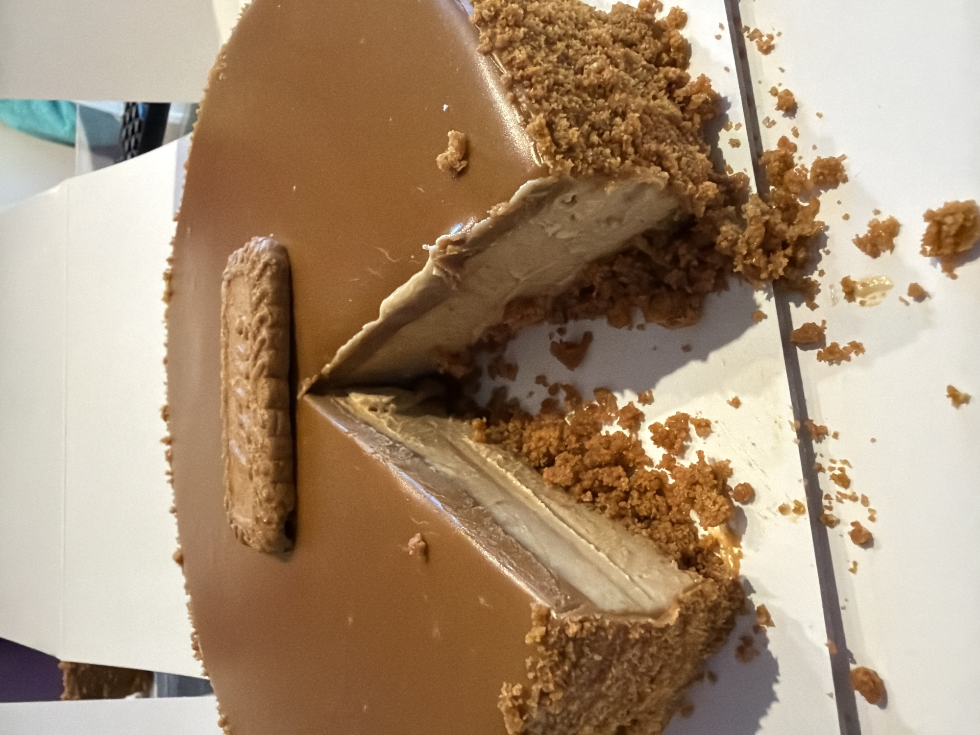 A whole, un-cut cake with a slice removed is shown in the image. The cake has a smooth, caramel-colored topping and a crumbly, brown crust around the sides. A single biscuit is placed on top of the cake. The cake is on a white surface, possibly a table or cake board. In the background, white cardboard boxes and a teal object are visible, suggesting the cake might be presented or packaged. The image appears to be taken indoors, with natural light.