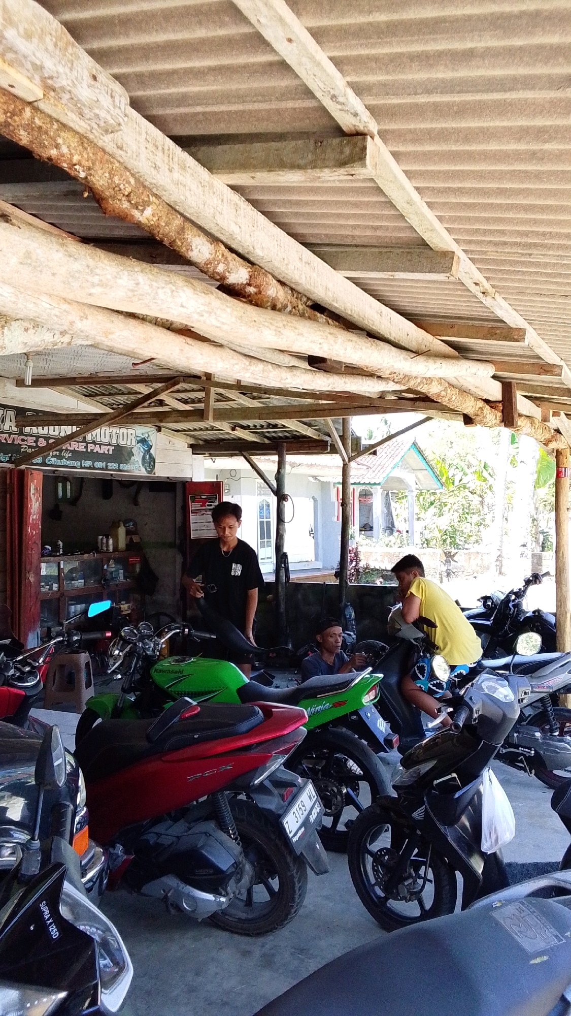 The image shows a motorcycle repair shop located in Cintaratu, Indonesia. The shop has a corrugated tin roof supported by rough-hewn wooden beams. Inside, several motorcycles are parked. In the foreground, a red scooter and a black motorcycle are visible. In the middle ground, a green Kawasaki Ninja motorcycle is parked next to a person in a black t-shirt holding a motorcycle part. Further back, another person in a yellow shirt is leaning on a motorcycle. The shop appears to be open-air with daylight filtering through the roof. Shelves with various items are visible in the background, along with a white building and trees outside. The overall impression is of a busy, functional workshop.
