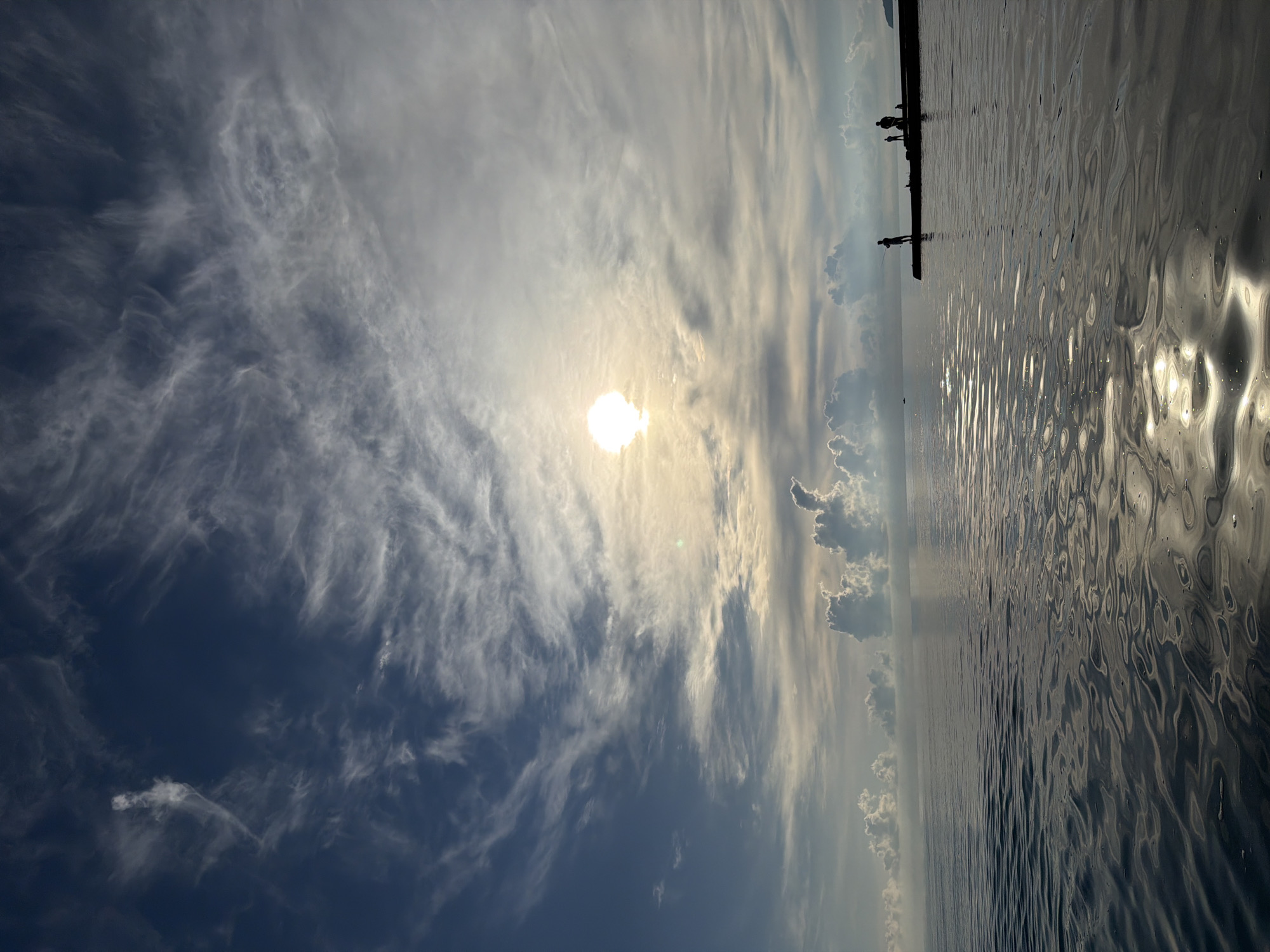 This media file captures a scenic view of the sky and water at Dederkoy, Russia. The image is oriented vertically, with the sky occupying the left half and the water the right.
The sky is dramatic, filled with wispy cirrus clouds that are illuminated by the bright, low sun, suggesting either sunrise or sunset. The sun itself is a prominent, glowing orb in the upper left quadrant, its light diffused by the clouds, creating a soft, ethereal glow. In the distance, silhouetted figures of people are visible standing on what appears to be a pier or breakwater. Some of the figures seem to be fishing, with their postures suggesting activity.
The water in the foreground is choppy, reflecting the sky and sunlight in shimmering patterns. The interplay of light and shadow on the water's surface creates a dynamic and almost abstract texture. The overall mood is serene and contemplative, with the vastness of the sky and the gentle movement of the water creating a sense of peace. The location cues point to Dederkoy, Russia, and the time of day is likely early morning or late evening due to the low sun angle and dramatic cloud illumination. There is no visible text in the image.