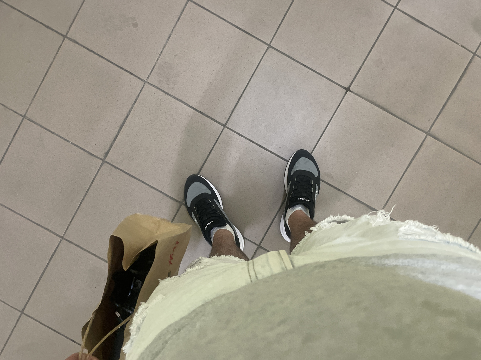 The image shows a close-up, high-angle shot of a person's legs and feet wearing black and gray athletic shoes with white soles and socks, standing on a tiled floor. The person is also wearing light-colored, distressed denim shorts. A brown paper shopping bag is visible to the left, with what appears to be a red logo. The floor is made of square, beige tiles with dark grout lines. The scene is likely indoors, possibly in a commercial establishment or a home. No specific landmarks of Kyiv, Ukraine, are identifiable in the image.