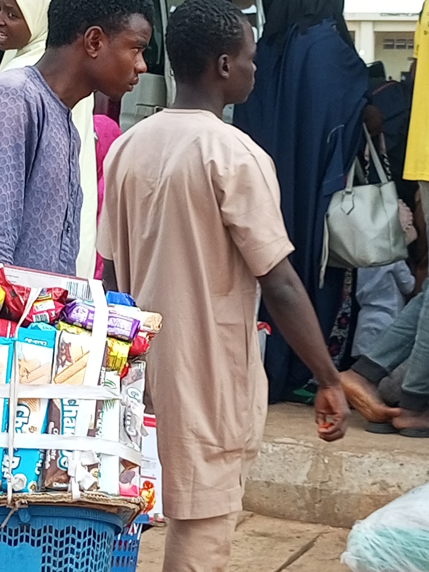 In Gombe, Nigeria, a young man stands in the foreground, wearing a light brown, knee-length tunic with short sleeves and a high neckline. He is facing away from the camera and to the right, with his left arm extended downwards and his hand reaching towards a small red object on the ground. His hair is dark and curly.
To his left, another young man is partially visible, wearing a long-sleeved, light purple tunic with an intricate patterned design. He is looking towards the right, his head turned slightly. Further to the left, a woman wearing a light yellow headscarf and a white garment is visible, with her mouth open as if speaking.
Behind the central figure, a woman in a dark blue, flowing garment is visible, carrying a light gray handbag over her right shoulder. To her right, a person wearing a yellow t-shirt and a dark garment is partially visible. Several other individuals are in the background, their features obscured.
In the immediate foreground, a blue plastic basket is filled with various packaged snacks, including bars of what appear to be biscuits and wafers, some with colorful wrappers. The basket is on the ground, and the snacks are stacked high within it. The ground appears to be a mixture of concrete and dirt.