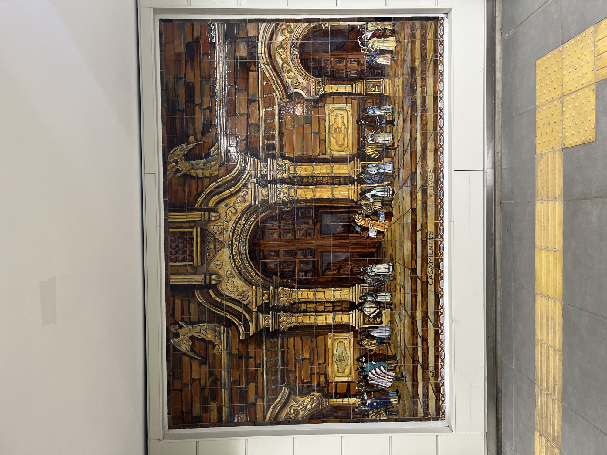 The image shows a tile mural depicting a colonial-era scene. It is framed in white and appears to be a wall decoration. The mural is rendered in brown and gold tones, depicting a building façade with figures dressed in historical attire. The tilework has a slightly aged appearance. The side of the tile mural reads "CASAMENTOS 1939" in brown tones, with a brown and tan tiled geometric pattern lining the border. Next to the white trim that surrounds the mural, there is a gray floor with a long yellow tile strip.
The location is likely a subway station in Buenos Aires, Argentina, as these tile murals are a feature of the city's underground transportation system, the Subte.