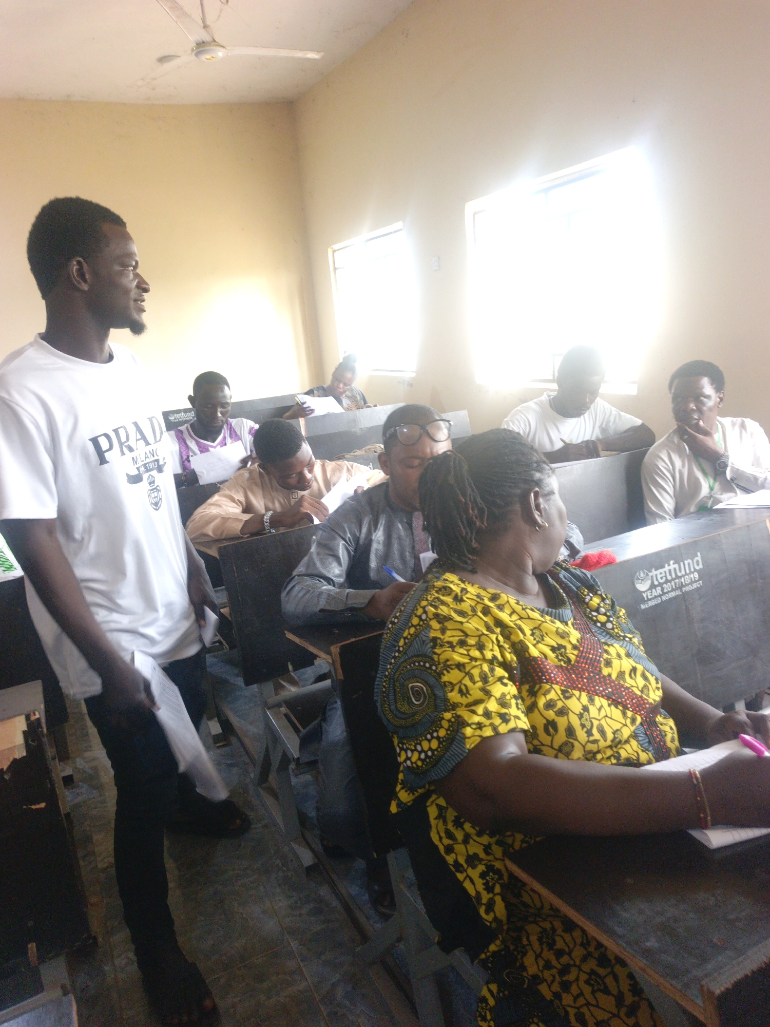 The image depicts an indoor scene, likely a classroom, in the city of Unknown, Unknown. There are multiple individuals present. A man stands in the foreground, wearing a white t-shirt with "PRADA MILANO" printed on it and dark pants. He appears to be holding papers. Seated in rows behind him are several other people, most of whom are looking down at papers on their desks, appearing to be writing. One woman is visible in the foreground, wearing a yellow and red patterned dress, also writing on paper. The desks have dark surfaces. The room has light-colored walls and two windows that are emitting bright light. A ceiling fan is visible on the ceiling.