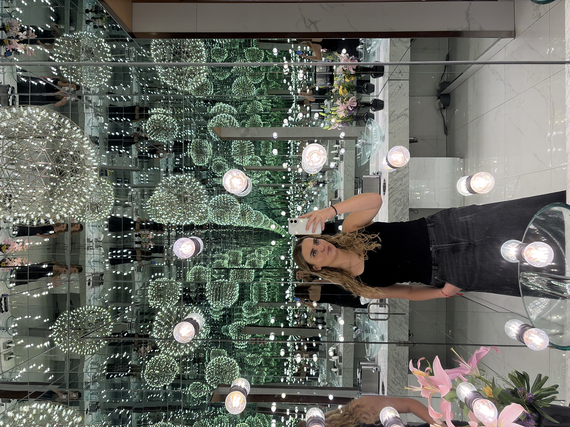 A medium shot presents a fair-skinned, blonde woman taking a selfie with a smartphone. She is dressed in a black t-shirt and gray denim pants. The surrounding environment appears to be a bathroom, enhanced by mirrors that create an infinite reflection of chandeliers and light fixtures. The walls and floor are decorated with a white and gray marble pattern. The scene's location is Unknown, Unknown.