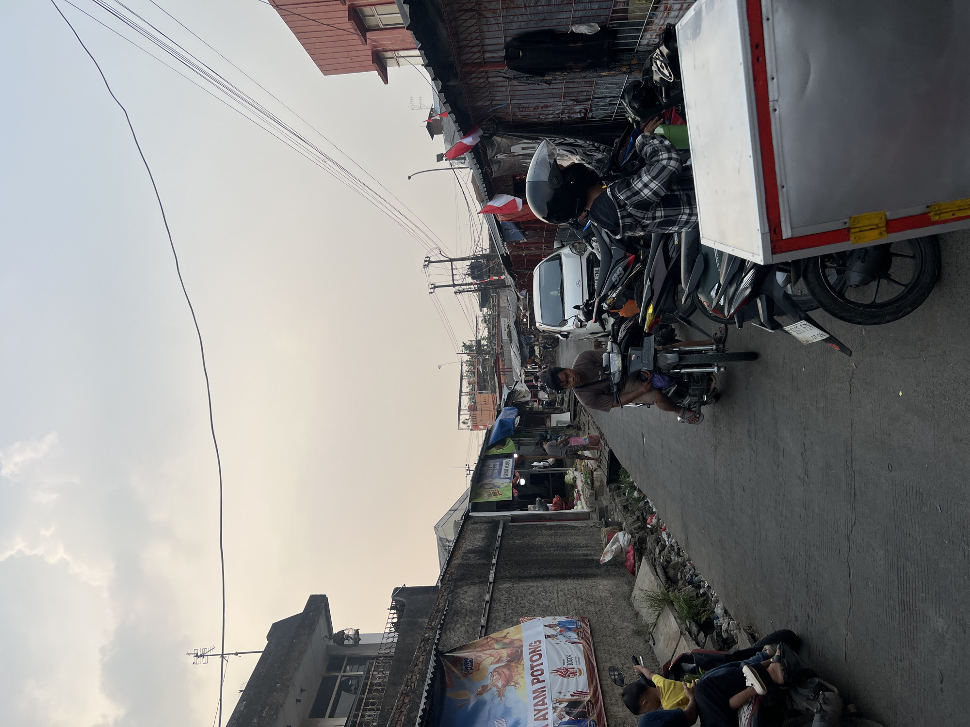 The image depicts a street scene in Citeureup, Indonesia, likely a market area. The sky is overcast with pale clouds. The street is paved with concrete and is lined with buildings and market stalls on both sides.
On the left side, a concrete wall supports a building above. A large banner with Indonesian text, including "AYAM POTONG" (cut chicken) and "BOGOR," is affixed to the wall. Several people are visible in the foreground and mid-ground. A child in a yellow shirt is sitting on a motorcycle on the left, pointing towards the right. Another person is seated behind the child on the same motorcycle. Further down the street on the left, a person is walking away from the camera.
The right side of the street features market stalls with awnings and merchandise. Several Indonesian flags are displayed on poles above the stalls. Various motorcycles are parked along the street. In the mid-ground, a white car is parked. Two men are seated on motorcycles, one in a plaid shirt and helmet, the other in a brown shirt. A large, silver insulated container with red and yellow trim is positioned in the foreground on the right. The overall impression is a bustling, somewhat cluttered urban environment.