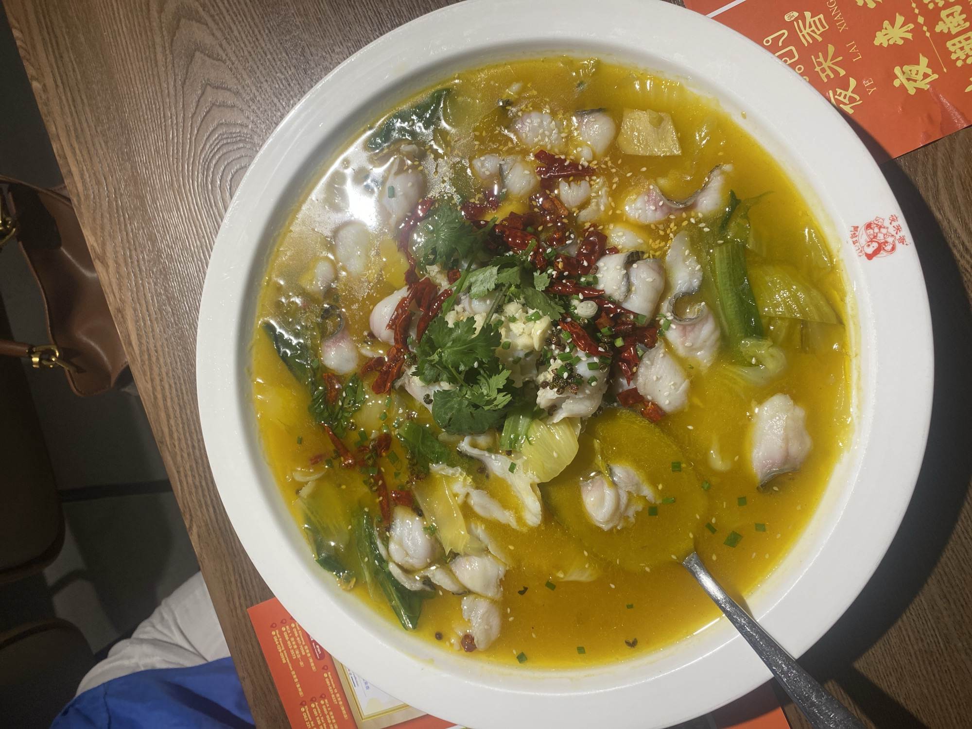 The primary subject is a large, oval white bowl filled with a vibrant yellow-orange soup, prominently featuring numerous slices of white fish fillet. The dish is generously garnished with dried red chili peppers, chopped green onions, white sesame seeds, and fresh cilantro, alongside green leafy vegetables submerged in the broth. A silver spoon rests in the soup.
The bowl is placed on a wooden table with a distinct grain pattern. In the background, a person wearing a blue and white shirt is partially visible, along with a brown leather bag featuring gold-toned accents and the edges of dark chairs.
The dish appears to be a spicy fish soup, likely a Hunan or Szechuan style given the abundant red chilies and bright, flavorful broth. The setting suggests an indoor restaurant environment.
Visible text on a red menu card next to the bowl includes Chinese characters such as "夜来香" (Yè lái xiāng), which means "Night Fragrance," and "湖南" (Húnán), explicitly indicating Hunan cuisine. The text "YỄ LẠI XIANG" and "Khaosan Rd" is also visible, confirming the location as Bangkok, Thailand, and suggesting a Hunan restaurant in or near the famous Khaosan Road area. Several phone numbers are also listed on the card.
