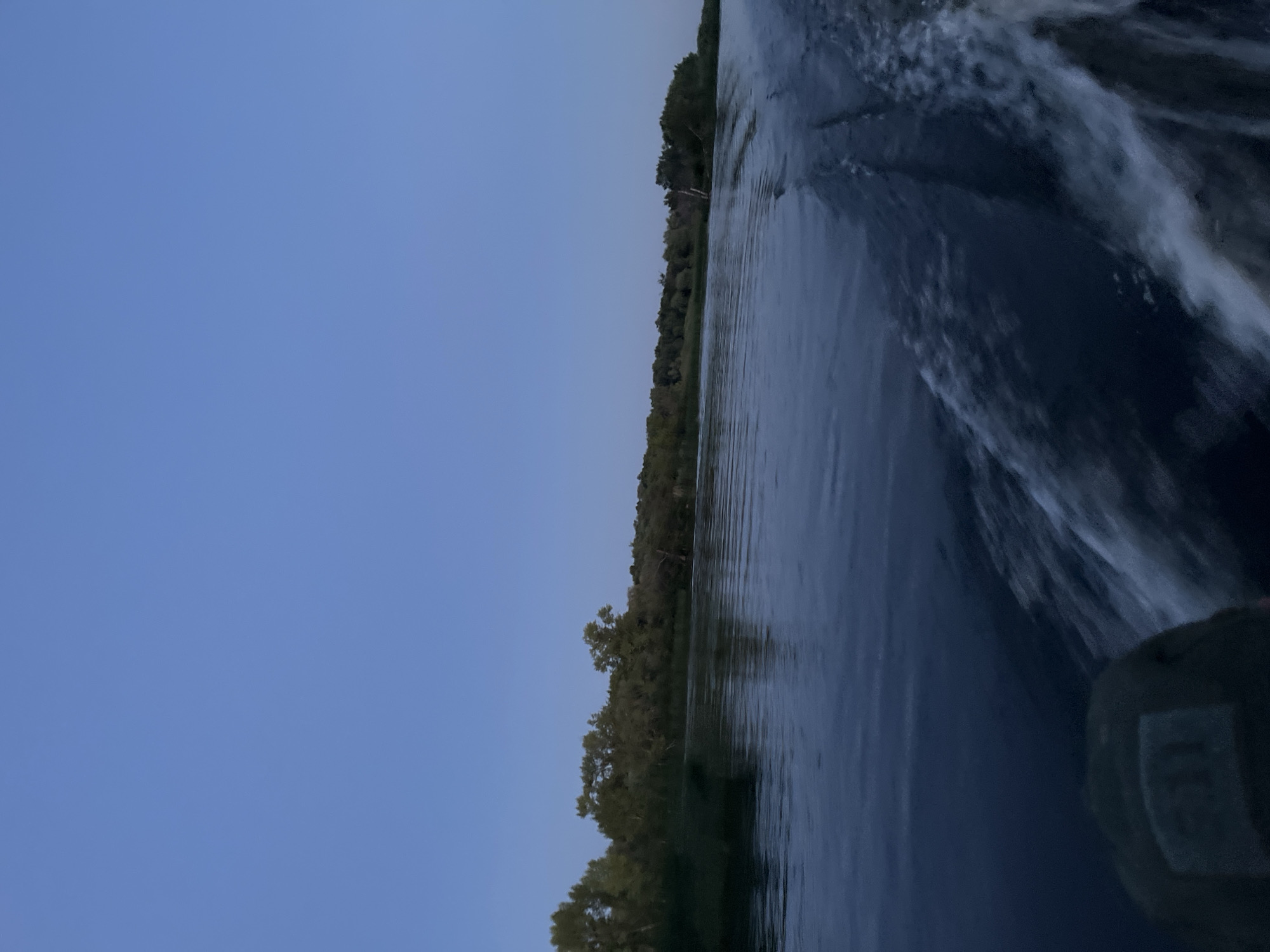 The image depicts a wide river at dusk, viewed from a moving boat. The sky is a clear, deep blue, transitioning to lighter shades near the horizon. On the left bank of the river, a dense line of trees forms a dark silhouette against the sky. The water is dark and reflects the dim light, with visible ripples and a white wake trailing behind the boat on the right side. A portion of the boat's interior, including what appears to be a dark-colored seat with a textured surface, is visible in the lower left corner of the frame. The scene is tranquil, capturing a moment of travel along the Volga River, likely near Saratov, Russia, given the characteristic landscape of the region.