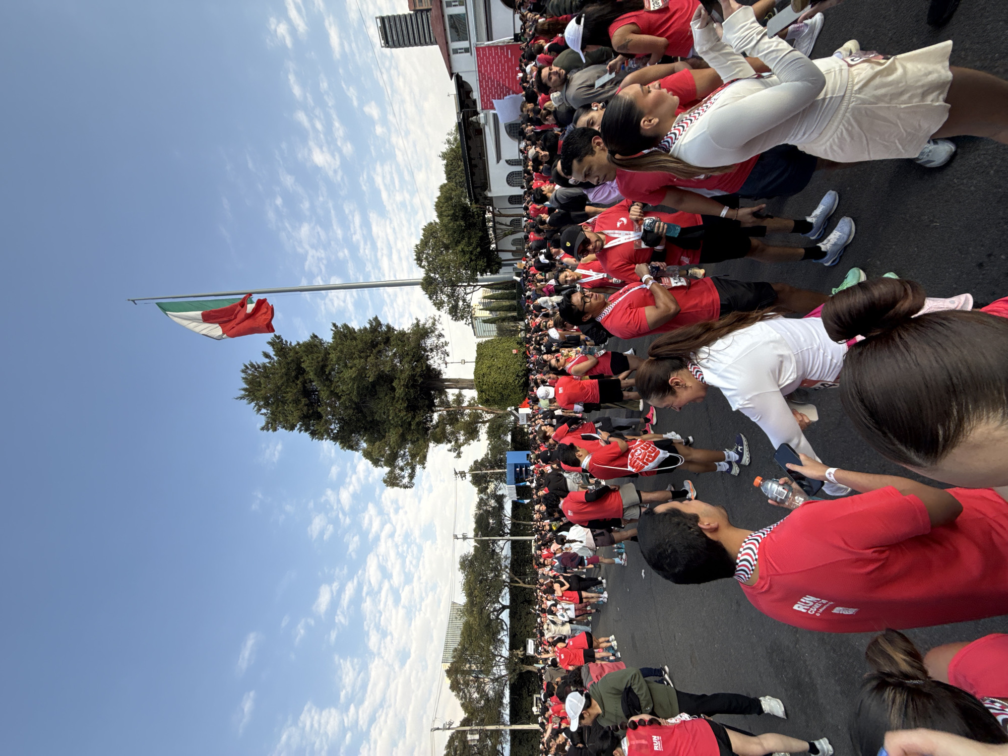The image shows a large crowd of people gathered outdoors, likely in Mexico City, Mexico. Most people are wearing red shirts with “RUN CDMX” printed on them. The crowd extends along a street lined with trees and buildings. The flag of Mexico is visible on a pole next to a tree. The sky is blue with scattered clouds. Some people are holding water bottles and phones. The event seems to be a running race or a similar gathering.