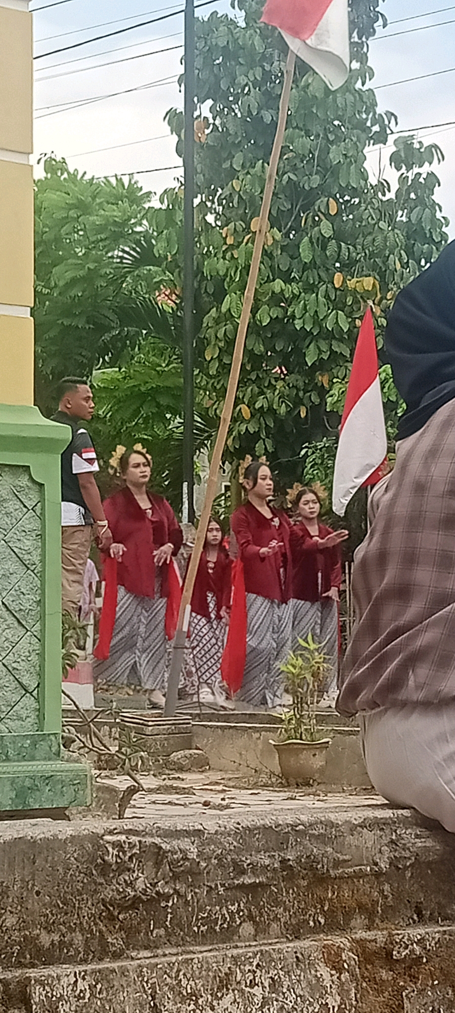 In Singgahan, Indonesia, a group of individuals is gathered outdoors, possibly for a cultural event or ceremony. A man in a black shirt with white and red trim stands to the left, facing away from the camera. In front of him, four women are arranged, dressed in traditional red blouses and patterned skirts. They appear to be performing or participating in a ritual, with their hands raised in a synchronized gesture. One younger girl, also in similar attire, stands slightly behind the group.
Two Indonesian flags, one red and white, are prominently displayed, attached to a tall wooden pole. The flags are positioned in the background, amidst lush green trees and foliage. The ground in the foreground is a rough, textured concrete surface, possibly a walkway or a platform. A potted plant sits on this surface, adding a touch of greenery to the scene. The overall impression is of a community event taking place in a well-maintained outdoor space.