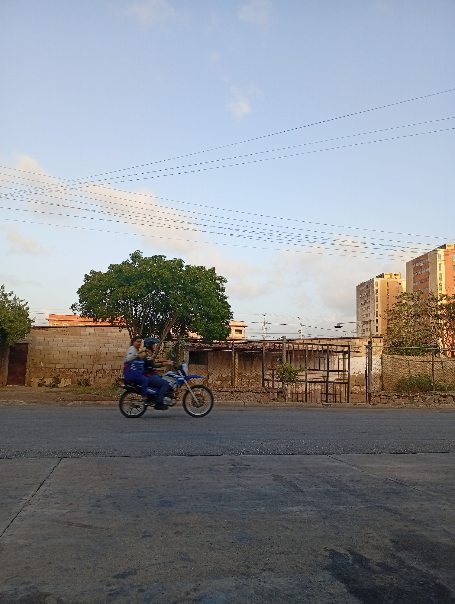 This image presents an outdoor street scene in Juan Griego, Venezuela, under a pale blue sky with scattered white clouds. The central activity features two people riding a blue and white dirt bike on a paved road, captured with motion blur, indicating movement from left to right. The driver is wearing a helmet, while the passenger, seated behind, looks to the right with a slight smile and is without a helmet.
The background is rich with urban elements. Behind the motorcycle, a long, light-colored concrete block wall runs parallel to the street, partially fronting a large, leafy green tree. To the right, various structures include a black metal gate leading into a property, and beyond that, several multi-story buildings dominate the horizon. Some of these buildings have a distinctive reddish-orange hue, suggesting they are catching the low-angle light of either early morning or late afternoon. Numerous utility lines stretch horizontally across the sky, crisscrossing in places. The foreground consists of a dark, asphalt road, with a strip of dirt and sparse vegetation separating it from the walls and fences. No visible text can be deciphered in the image. The overall impression is a candid glimpse into daily life in a residential or mixed-use urban area.