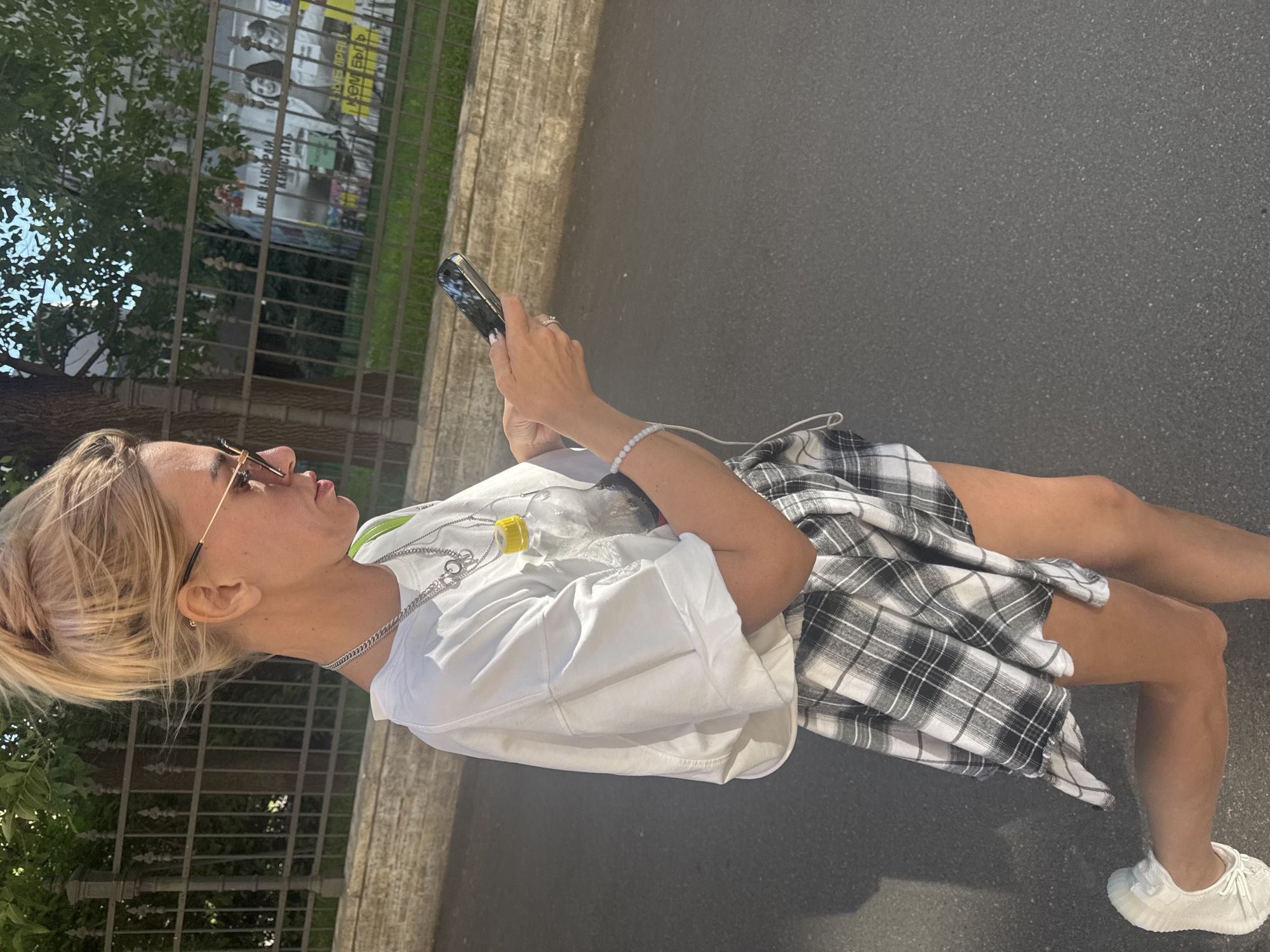 A young woman with blonde hair, wearing sunglasses, a white t-shirt, and a plaid skirt is looking at her phone while standing on a sidewalk. She is wearing a silver chain necklace and a light blue beaded bracelet. Her phone is black and appears to be an iPhone. In the background, there is a metal fence with posters behind it, and trees with green leaves. The ground is asphalt. The time of day appears to be daytime. The weather seems to be sunny. There is some visible text on the posters in the background, but it is not legible.