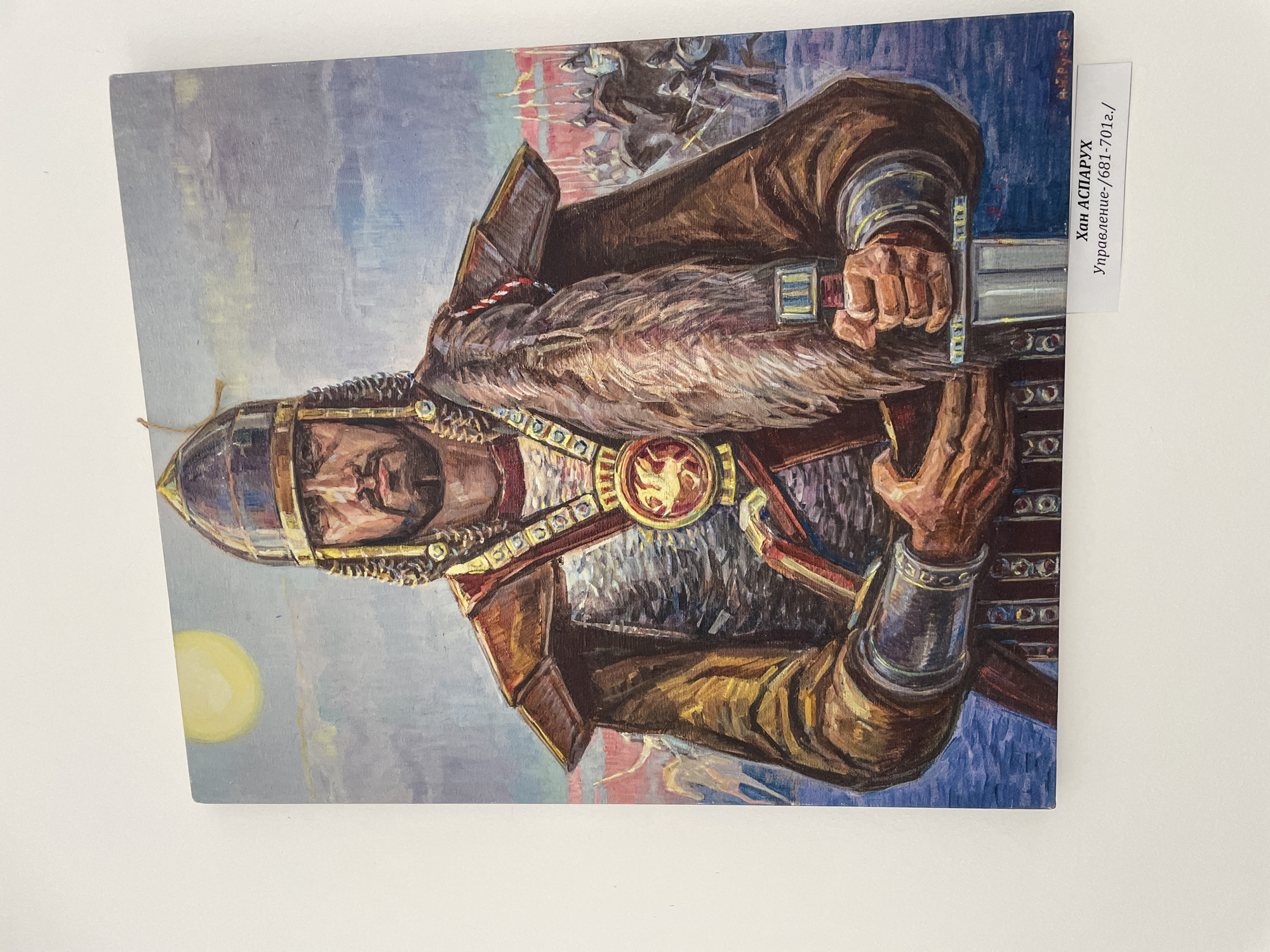 The image is a painting depicting Khan Asparuh, ruler of the First Bulgarian Empire. He is shown in armor with a helmet and a fur collar. He holds a sword with both hands. The background includes a sky with a yellow circle resembling the sun, and a scene of horses and figures in battle. A white label with the text "Хан АСПАРУХ Управление (681-701г.)" is affixed to the right side of the painting. The city of Zlatograd, Bulgaria, is not visible in the painting, but the painting is likely located there.