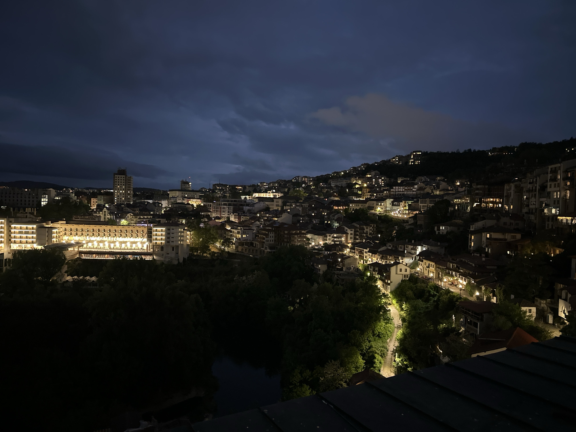 The image captures a nighttime view of Veliko Tarnovo, Bulgaria. The urban landscape is spread across a hillside under a dark, cloudy sky. Illuminated buildings and streetlights punctuate the darkness. A large hotel complex is visible on the left, emitting bright light. In the foreground, the tops of trees and a small body of water are seen, indicating a forested area or park. The image is taken from an elevated perspective, possibly from a building rooftop.