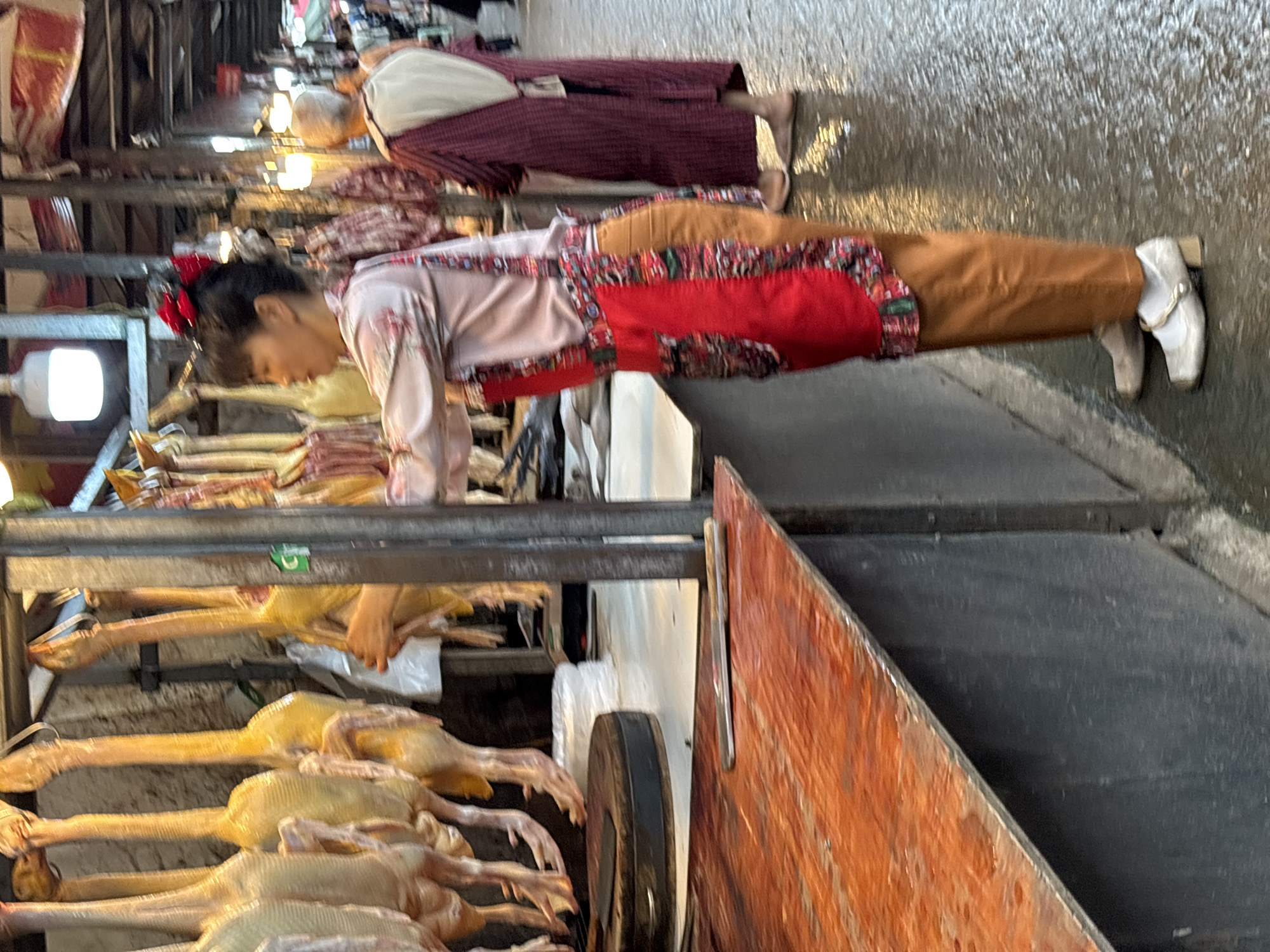 In a bustling market in Jiangjin, China, a woman wearing a light pink, floral-patterned top, red apron, and brown pants, inspects a rack of plucked ducks. She has a red hair accessory. The ducks are pale yellow and hang on metal racks. Another person in a burgundy outfit is further down the aisle. The ground is wet and reflective, suggesting a recent cleaning or damp weather. A wooden cart with a black wheel is in the foreground, adding to the market's cramped atmosphere.