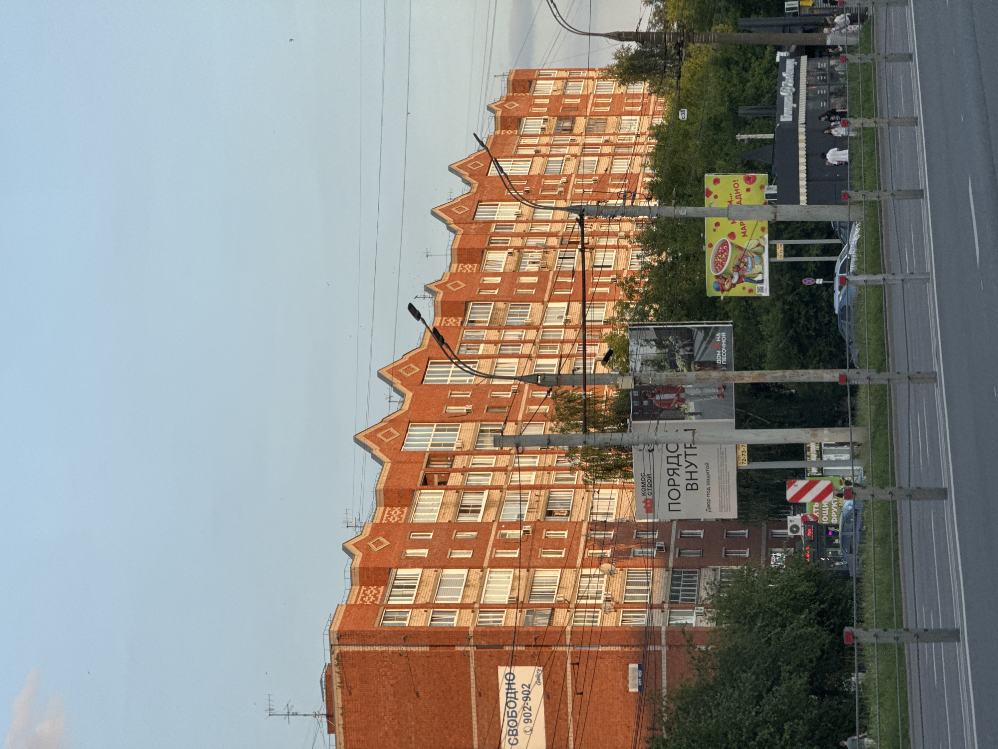 The image shows a multi-story brick apartment building with a distinctive stepped roofline. The building is illuminated by the warm light of the setting sun. In front of the building, there are several billboards, power lines, and streetlights. A road with cars and pedestrians is visible in the foreground. The building is located in Izhevsk, Russia, as indicated by a sign with the text "ИЖЕВСК" (Izhevsk) on it. The overall scene depicts a typical urban environment in Russia during the evening.