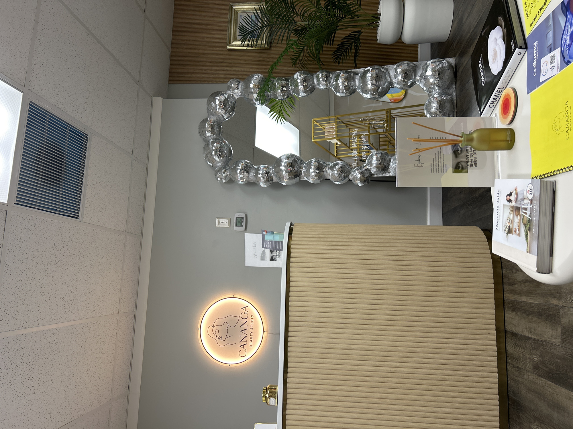 The image shows the interior of a beauty studio, likely in Hallandale Beach, United States of America. The studio features a light wood reception desk with vertical ribbed detailing and a brass base. Above the desk, a circular illuminated sign displays "CANANGA BEAUTY STUDIO" with a stylized graphic. To the right of the desk, a tall, arched mirror is adorned with numerous small disco balls. Further to the right, a potted palm plant sits in a white pot, next to a wall with vertical wood paneling. In the foreground, a white coffee table holds several items, including a book titled "Magnolia Table," a yellow notebook with "CANANGA BEAUTY STUDIO" printed on it, and a book with "CHANEL" on the cover. A small diffuser with reeds is placed on the coffee table. The overall environment appears clean and modern, suggesting a commercial space.