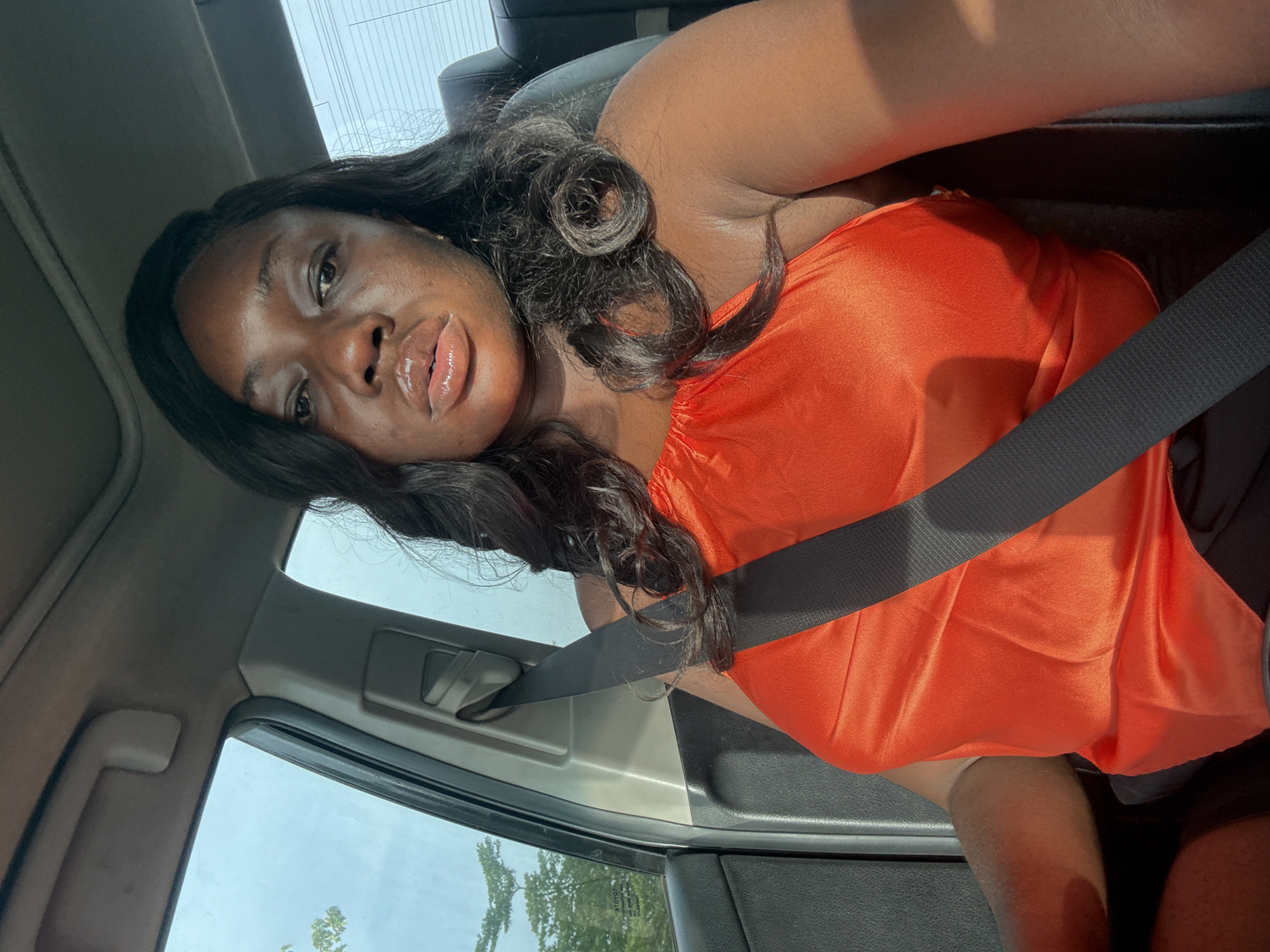 A medium, angled, indoor shot shows a dark-skinned woman in the passenger seat of a vehicle, secured with a dark-colored seatbelt. She is wearing a bright orange, off-the-shoulder top. Her hair is black and styled with loose waves and curls. The interior of the car is visible, including the door panel, window, and roof. Outside the car window, green foliage and possibly power lines can be seen, suggesting an outdoor setting. The woman's facial expression is relaxed, and her lips appear glossy.
I have no information about the location of the photo, so the location provided cannot be verified.