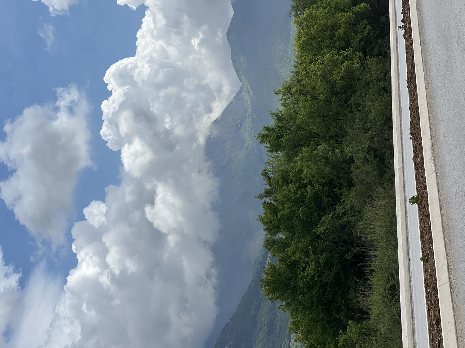 This image, taken in Kavač, Montenegro, presents a vibrant mountainous landscape under a dynamic sky. The top portion of the frame is dominated by a bright blue sky, generously filled with large, volumetric white cumulus clouds, some appearing fluffy and others darker, hinting at atmospheric depth or a potential change in weather.
Below the sky, lush green mountains or hills stretch across the midground, their peaks and slopes softly veiled by a light mist or haze, suggesting a moist environment. A dense band of dark green trees and bushes forms a natural barrier in front of the mountains, showcasing the rich vegetation of the area.
In the foreground, a grey paved surface, likely a road or a wide path, runs horizontally along the bottom of the image. It is clearly defined by a white line and bordered by a narrow strip of earth, resembling a median or planter, where small, resilient green plants sprout from the soil. This strip is contained by light-colored concrete curbs.
The scene depicts a peaceful, expansive natural setting, likely captured during daytime, possibly late morning or afternoon, given the clear illumination. There is no human activity taking place, and no visible text is present. The overall impression is one of natural beauty and tranquility in a mountainous region.