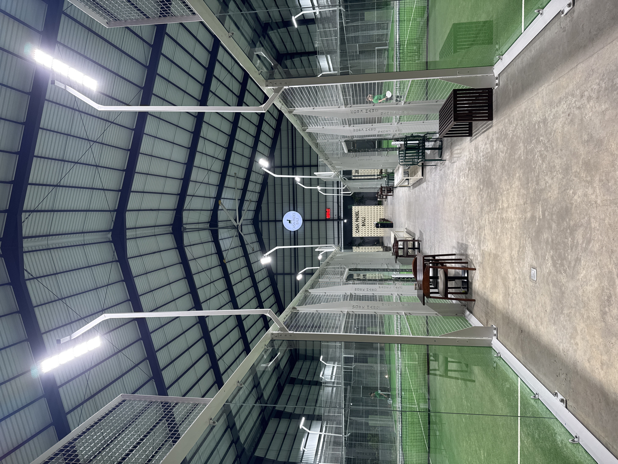 The image shows an indoor paddle tennis facility, identified as Casa Padel Bali, located in Kuta Selatan, Indonesia. The facility features multiple paddle tennis courts with green playing surfaces and glass or wire mesh enclosures. Overhead, a high metal roof structure supports lighting fixtures. Along the side of the courts are seating areas with various types of chairs and benches. A person is visible on one of the courts, suggesting the space is in use. The floor is a light-colored concrete.