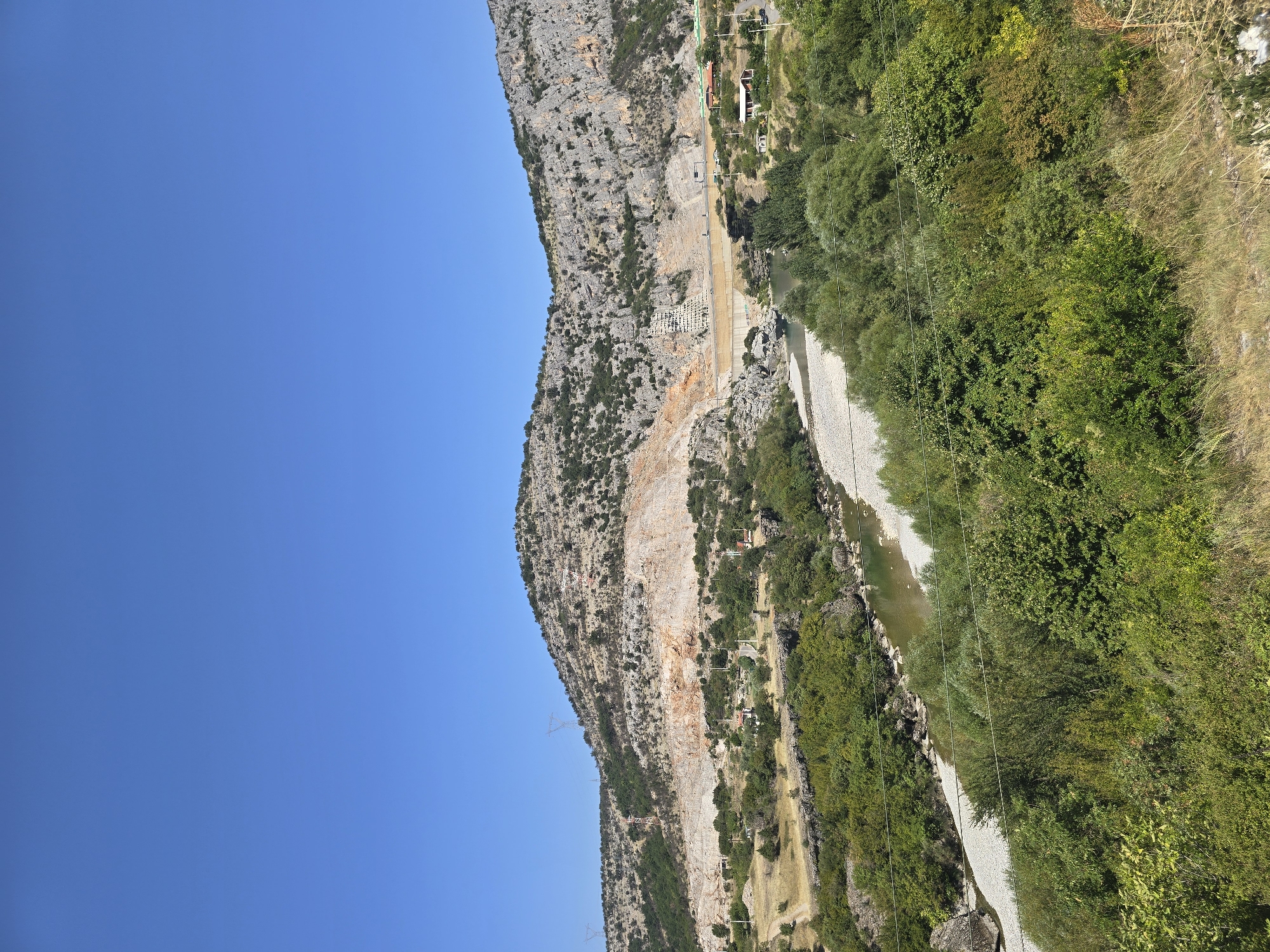 The image shows a panoramic view of a mountainous landscape in Podgorica, Montenegro, with a river winding through a valley. The foreground is dominated by lush green vegetation, including trees and shrubs, with dry grass and a partially visible fence on the right. A narrow, light-colored river runs through the middle ground, its banks lined with pebbles and rocks.
In the middle ground, a large dam structure is visible across the river, with construction work or some sort of scaffolding on its left side. The dam is built into the side of a steep, rocky mountain. The mountain itself is characterized by exposed rock faces, some with evidence of quarrying or erosion, and sparse vegetation. Several power lines and transmission towers are visible on the mountainside.
Further into the distance, the rugged terrain continues, with more rocky slopes and scattered trees. The sky is a clear, bright blue, indicating a sunny day. A few small buildings, possibly houses or auxiliary structures related to the dam, are scattered near the river and the base of the mountain. The overall scene suggests an area of natural beauty undergoing some form of human intervention or development.