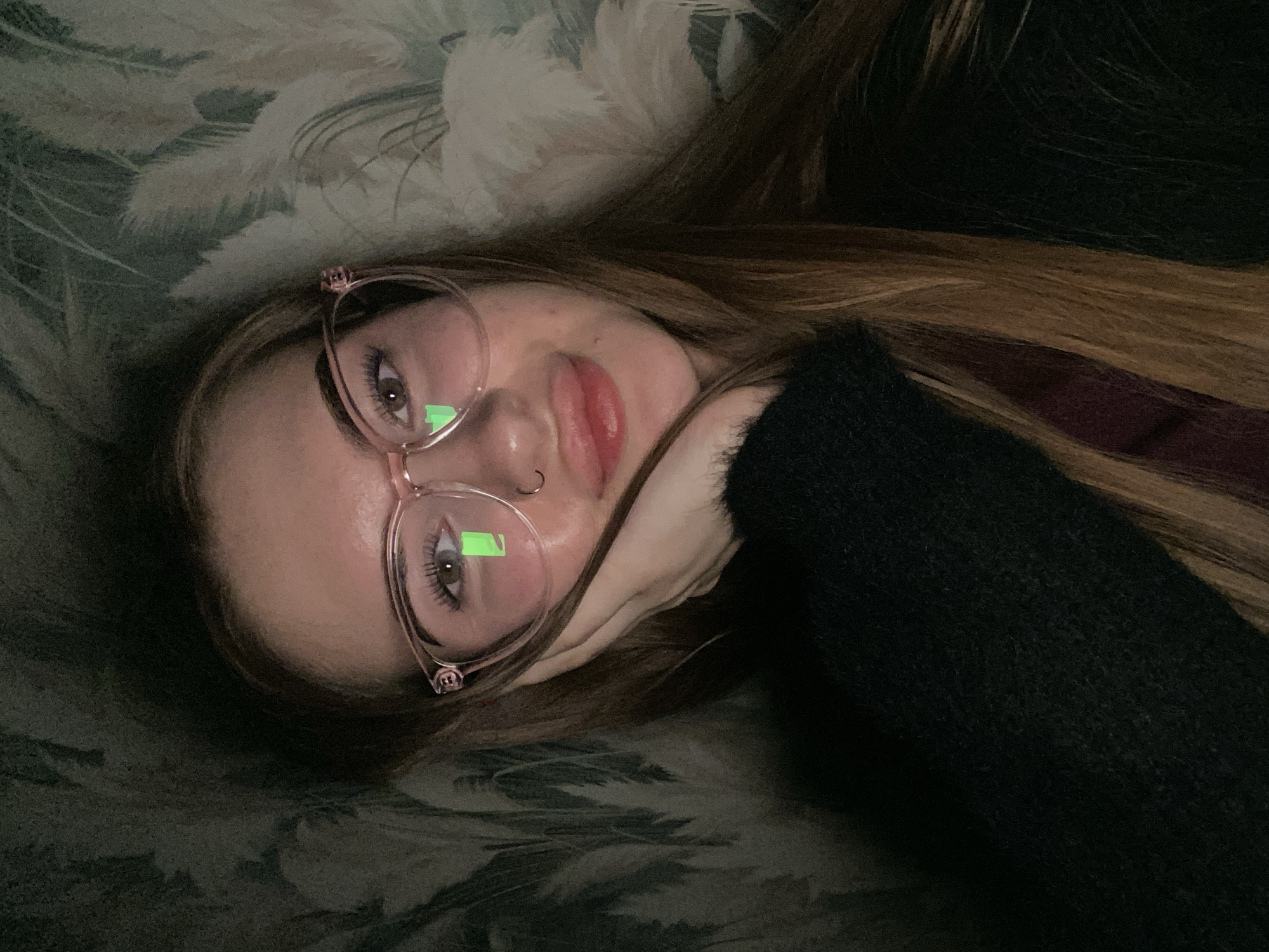 The image shows a person, likely female, lying down indoors and looking directly at the camera. Their head is tilted, and their left hand supports their chin or cheek. They have long, light brown hair and wear glasses with light pink, possibly translucent, frames. A small silver hoop nose ring is visible on their left nostril.
Notable details include prominent eyelashes and dark irises visible through the left lens. Bright green rectangular reflections are visible on both lenses of the glasses, suggesting an artificial light source, possibly a screen, in front of the subject. The person is wearing a dark, possibly black, fuzzy or knitted garment on their right arm and shoulder.
The background consists of a soft, patterned textile, likely upholstery or a blanket, featuring light, feathery or botanical designs against a muted, dark green-grey fabric. The overall lighting is dim, suggesting an indoor setting during evening or night. The activity appears to be resting or relaxing, with a neutral and calm expression. No text is visible in the image.