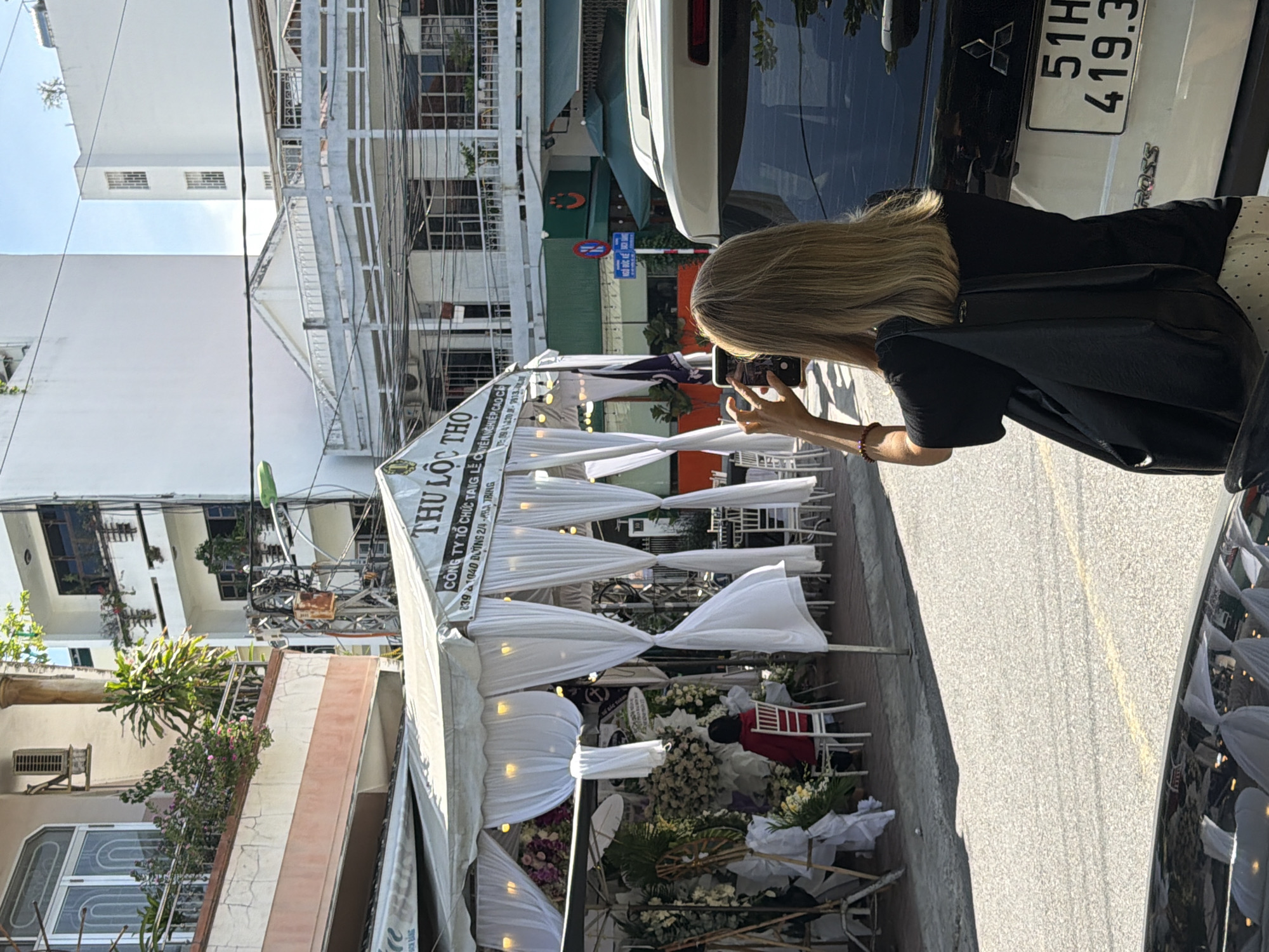 In Nha Trang Ward, Vietnam, a blonde-haired woman, seen from behind, stands on a paved road, holding a smartphone to take a photograph. She is wearing a black t-shirt, a black bag over her shoulder, and a white polka-dot skirt. To her right is the rear of a white SUV, identified by a license plate reading "51H 419.3". In the background, a white tent is set up, decorated with white drapery and floral arrangements, appearing to be an event venue. Inside the tent, a person in a red jacket sits on a white chair. The street scene is bordered by buildings and a blue sign with Vietnamese writing.