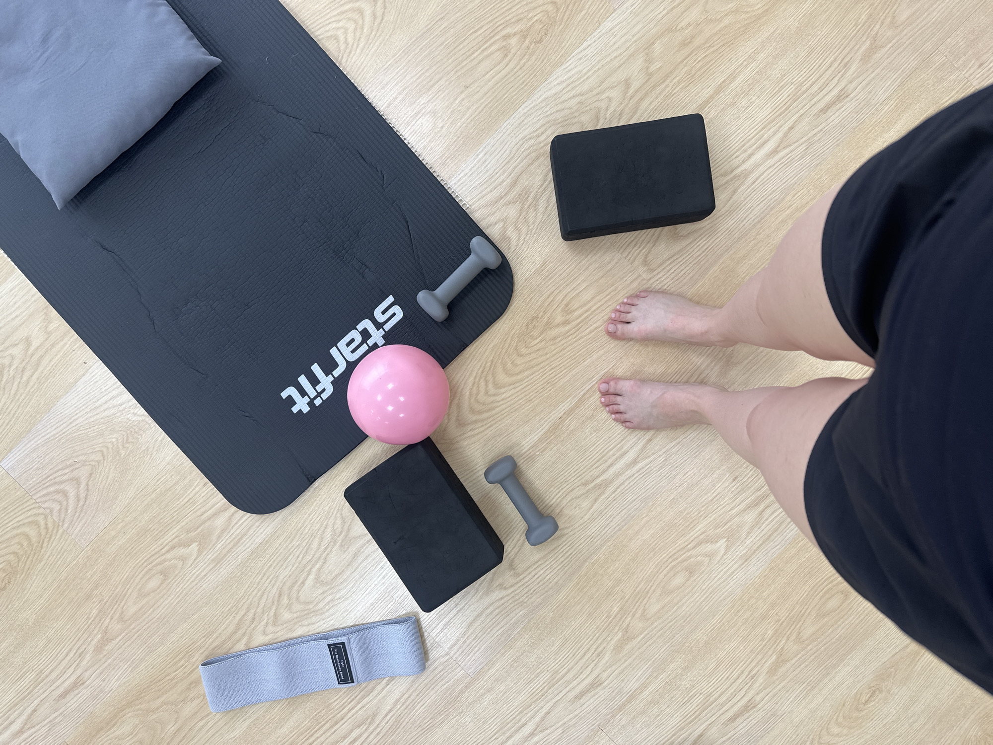 The image depicts a workout setting on a light wood floor. A black yoga mat with the word "starfit" in white lies diagonally across the frame with a grey pillow resting on the top left corner. A small pink exercise ball, a pair of light gray dumbbells, and two black yoga blocks are also arranged on the floor. A light gray resistance band is located at the bottom left corner of the frame. A person's feet and lower legs are visible to the right of the yoga mat, and the person is wearing a dark black garment that obscures the rest of their body. The setting is likely indoors, possibly a home gym or studio. There is no indication of the location being Votkinsk, Russia.