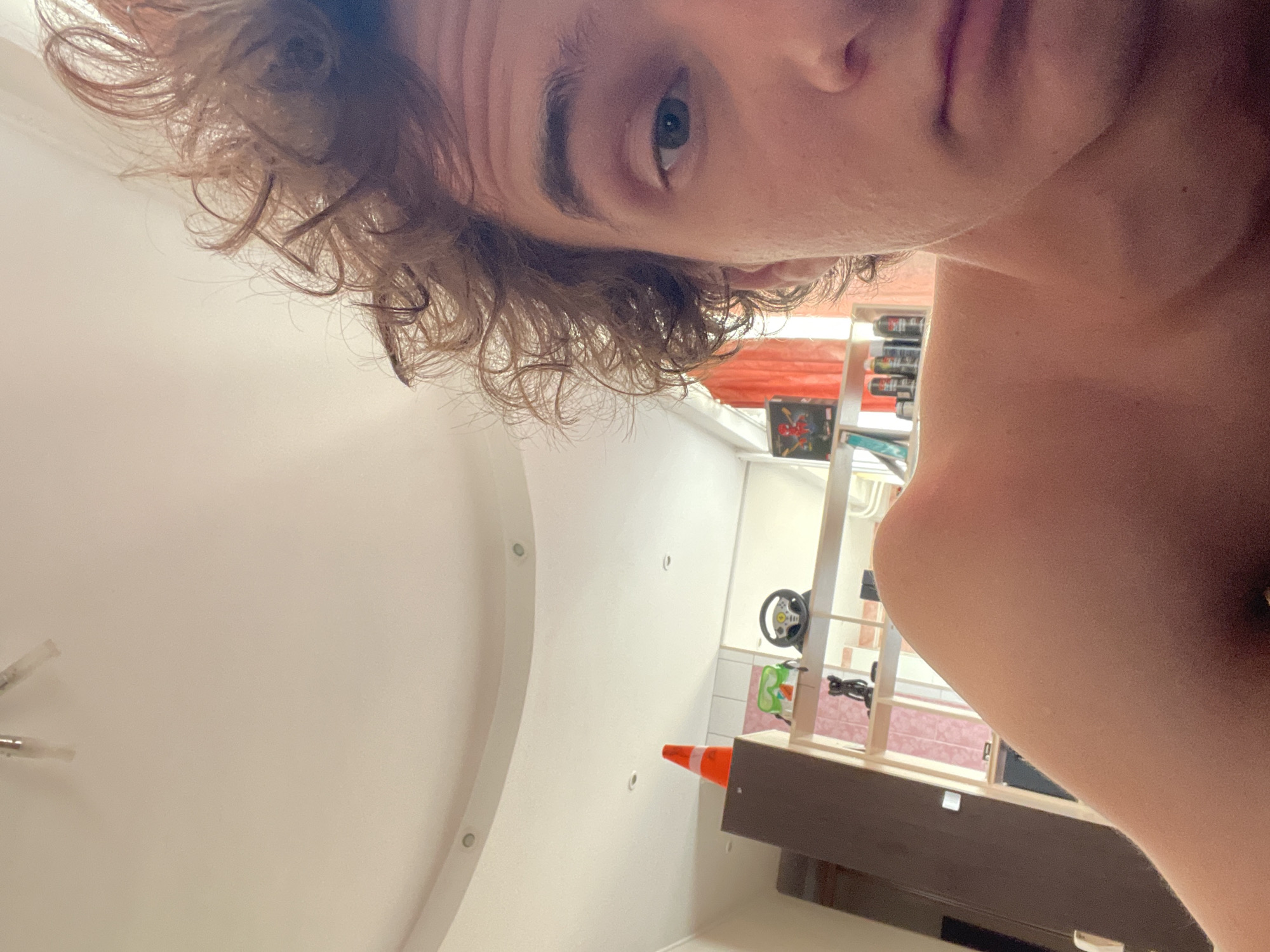 The image shows a close-up, selfie-style view of a young person's face and shoulder. They have curly brown hair and are looking towards the camera with a neutral expression. Part of their shoulder and upper chest is visible, and it appears flushed. In the background, a shelf unit is visible with several items, including what appears to be a toy steering wheel, a small figurine, and some spray cans. An orange traffic cone is also visible on top of a dark brown cabinet. The ceiling in the background is white and curved, with recessed lighting. The overall impression is of an indoor personal space, likely a bedroom or living area, but no specific details place it in Krasnoyarsk, Russia.