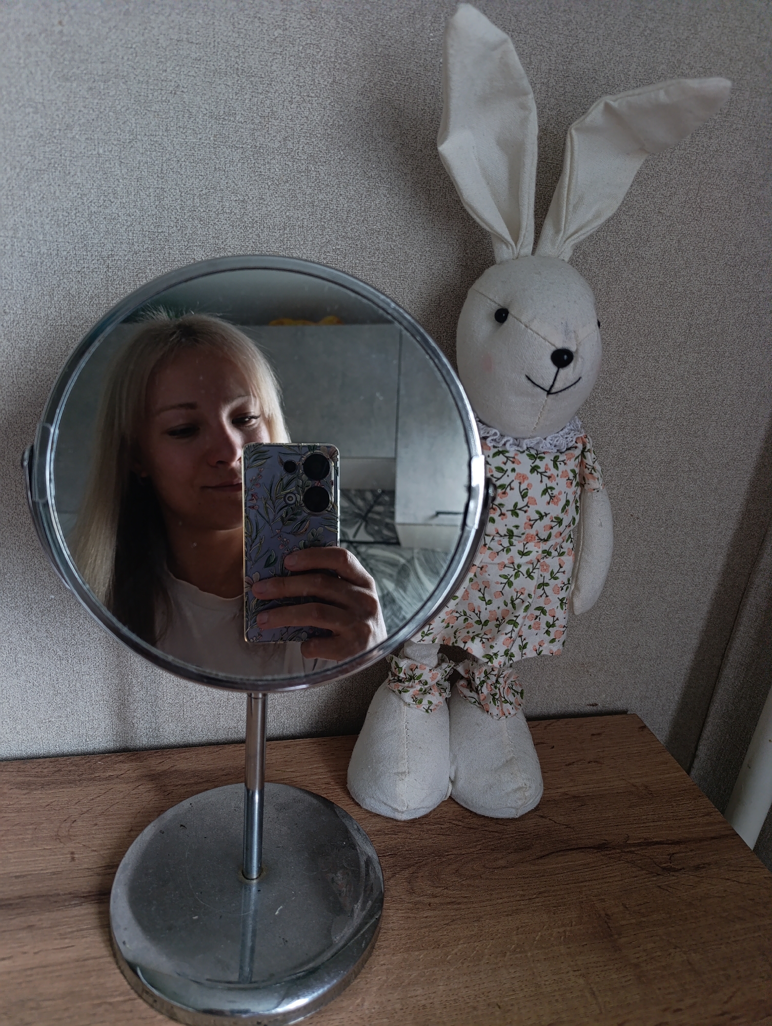 In a bright, indoor setting, a young woman with blonde hair takes a selfie in a circular mirror. She is holding a smartphone with a floral case, its screen displaying a colorful image. The mirror is attached to a silver stand with a circular base, resting on a wooden surface. To the right of the mirror, a plush white bunny toy stands dressed in a floral outfit. The bunny has long ears, black button eyes, and a stitched smile. Its dress features a pattern of small pink flowers and green leaves, and it also wears matching bloomers. The background consists of a textured, light-colored wall. The lighting suggests it is daytime. There is no visible text in the image. The overall mood is casual and personal.