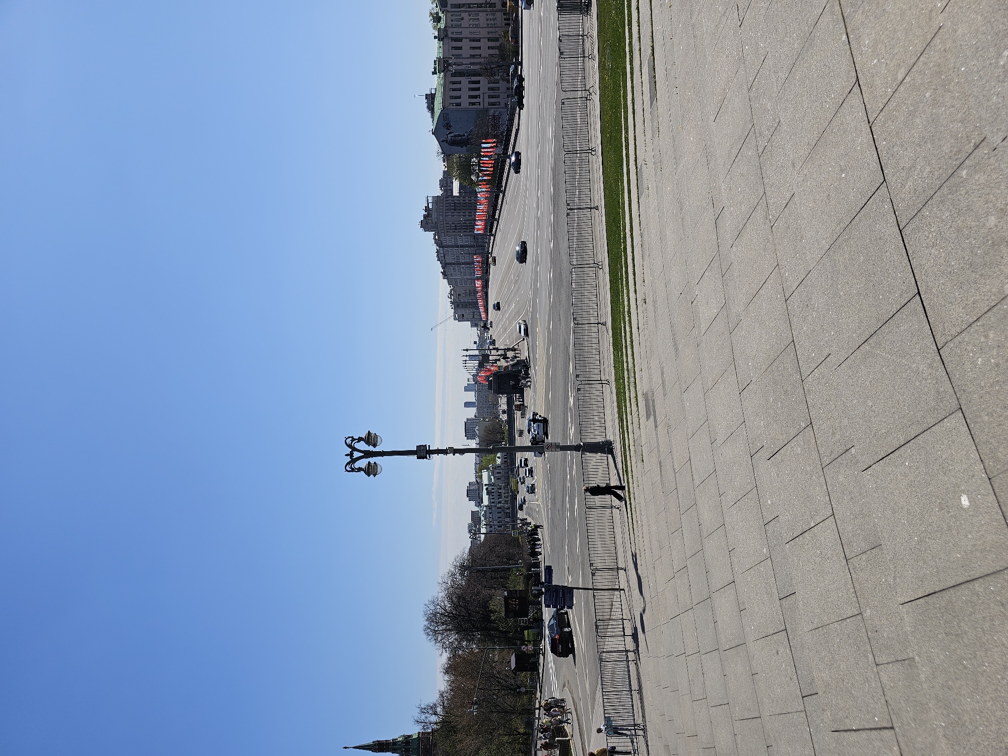 Here is a description of the image:
This image shows a street view from above in Moscow, Russia, likely taken on or near Red Square. The scene is oriented vertically, with a clear blue sky taking up a large portion of the left side of the frame. The right side of the image is dominated by a paved walkway or plaza, made of large, rectangular stones. Along the edge of the walkway, a metal barrier lines the road.
A major roadway stretches into the distance, flanked by buildings on both sides. Red banners are visible hanging from the buildings. Various vehicles, including cars and what appears to be military vehicles, are moving along the road. A few pedestrians are walking along the sidewalk near the barrier.
A decorative street lamp stands tall near the walkway. In the lower left, the distinctive spire of the Kremlin is partially visible, confirming the location.