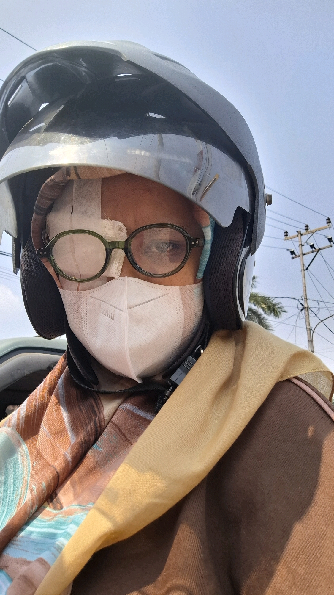 A person wearing a motorcycle helmet, glasses, and a face mask is shown from the chest up, with the image looking upwards towards the sky. The person's forehead and the area around their eyes are bandaged. The person is wearing a brown jacket and a patterned scarf draped around their neck and shoulders. The background shows a bright sky, power lines, and a utility pole, suggesting an outdoor urban environment. The scene appears to be captured in Bandar Lampung, Indonesia, given the presence of typical urban infrastructure and the style of the helmet and clothing, which are common in the region.